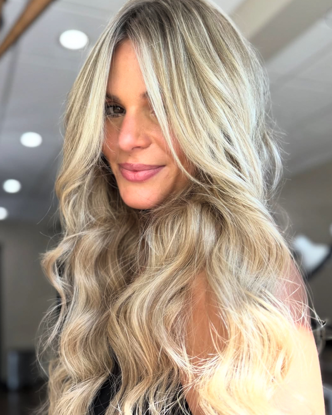 Close-up of a woman with long, wavy blonde hair and a subtle smile, looking over her shoulder in a well-lit indoor setting.