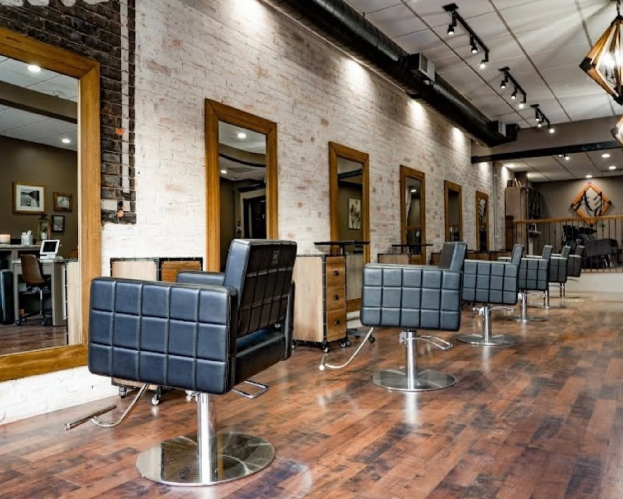 Interior of a modern hair salon with black styling chairs on a wooden floor, white brick wall, mirrors framed in wood, and ceiling track lighting.