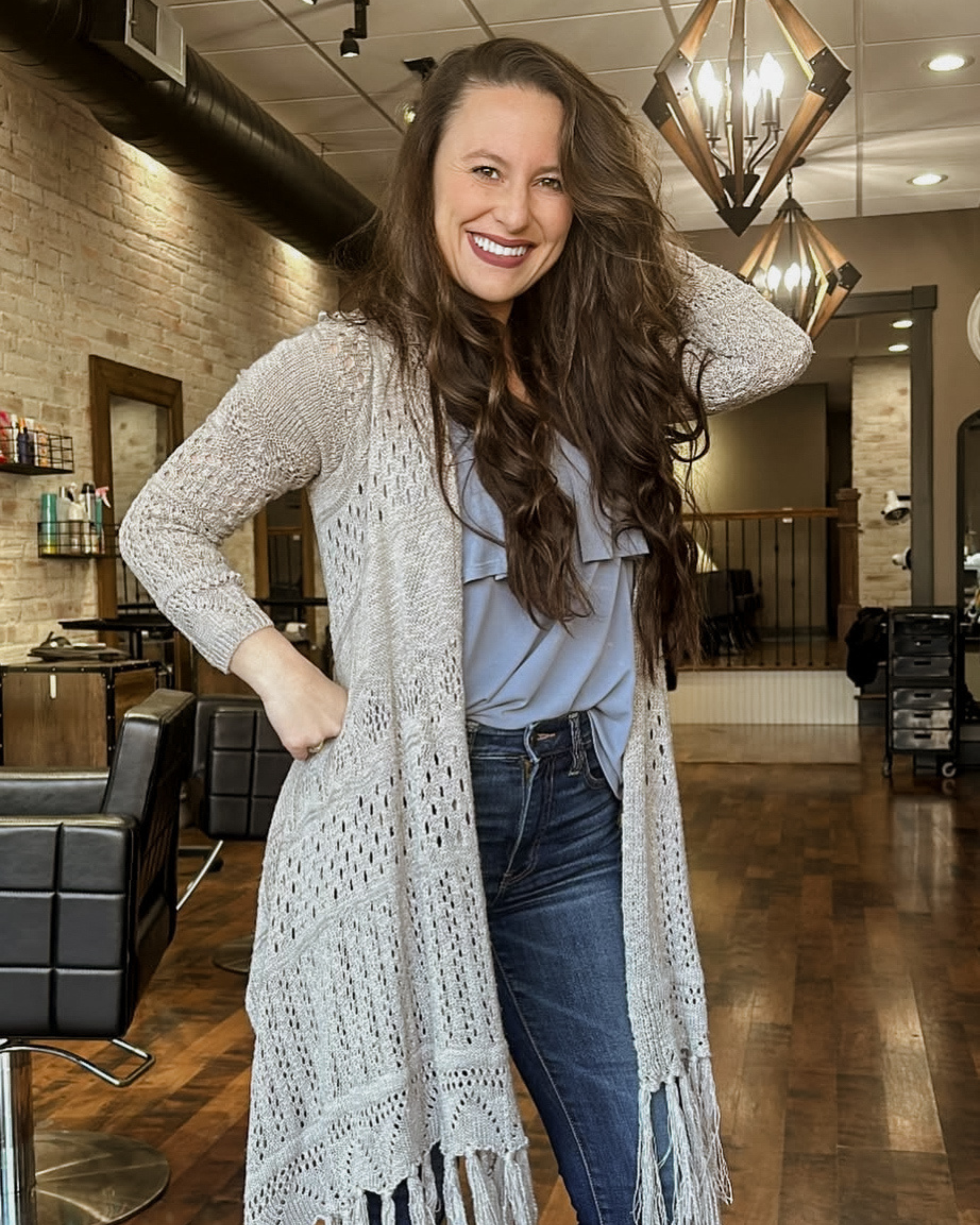 A woman with long, wavy brown hair smiling in a hair salon or similar space, wearing a gray shirt, jeans, and a long, knitted cardigan.
