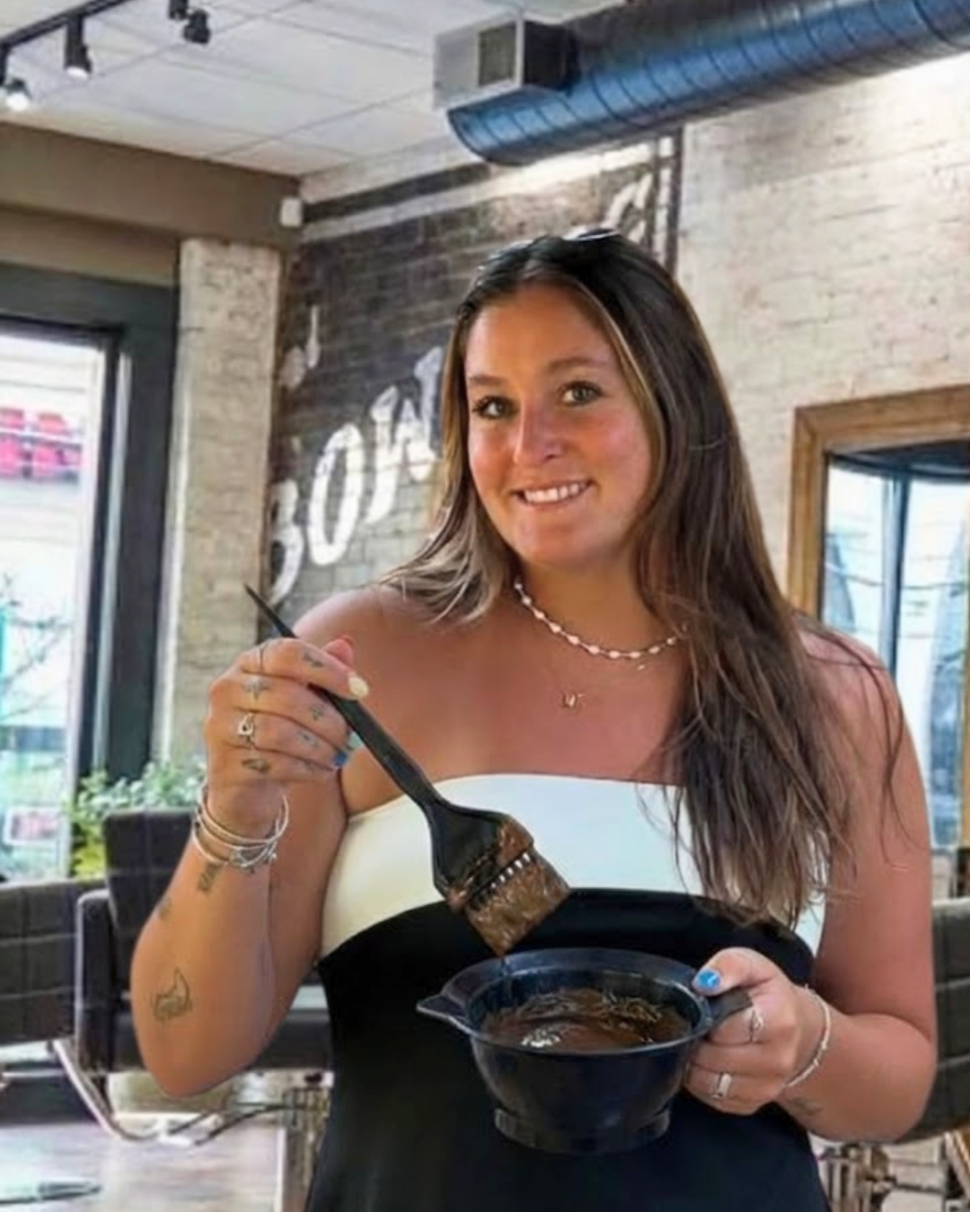 A woman with long brown hair in a cafe, holding a bowl of ramen with chopsticks, smiling at the camera.