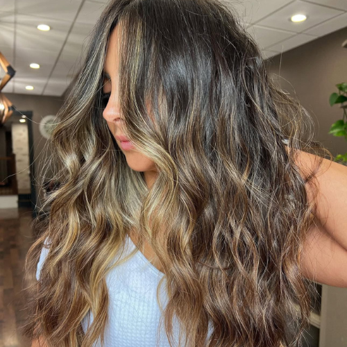 A woman with long, wavy hair with dark roots and blonde highlights, wearing a light-colored top, in an indoor setting.