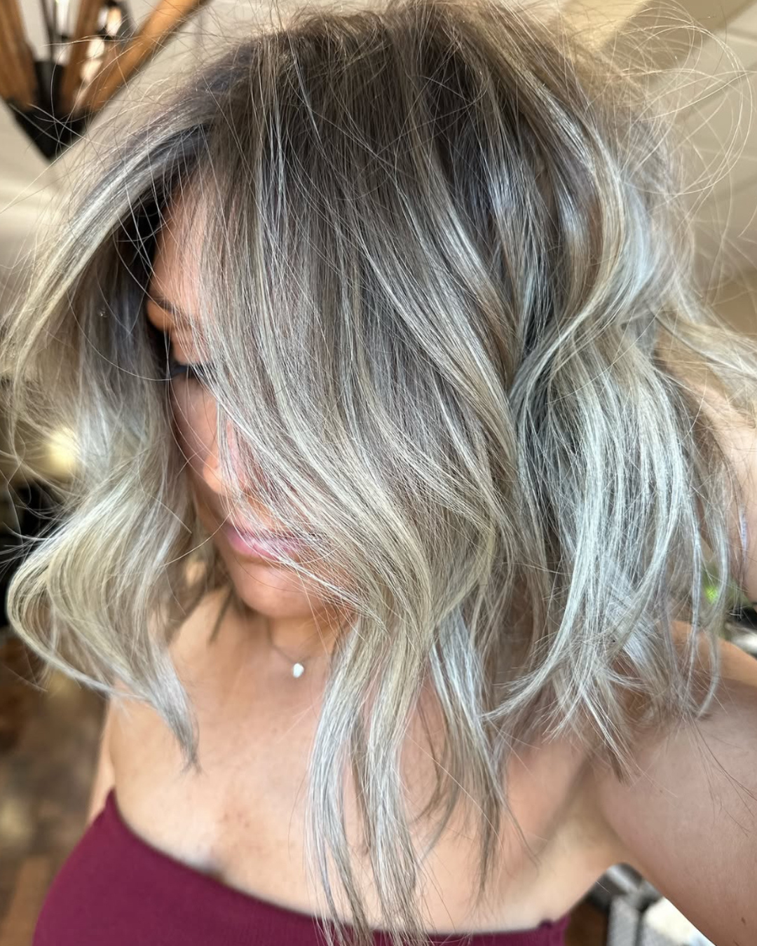 Close-up of a woman with shoulder-length wavy gray and blonde hair, partially covering her face, wearing a maroon top and a small necklace.