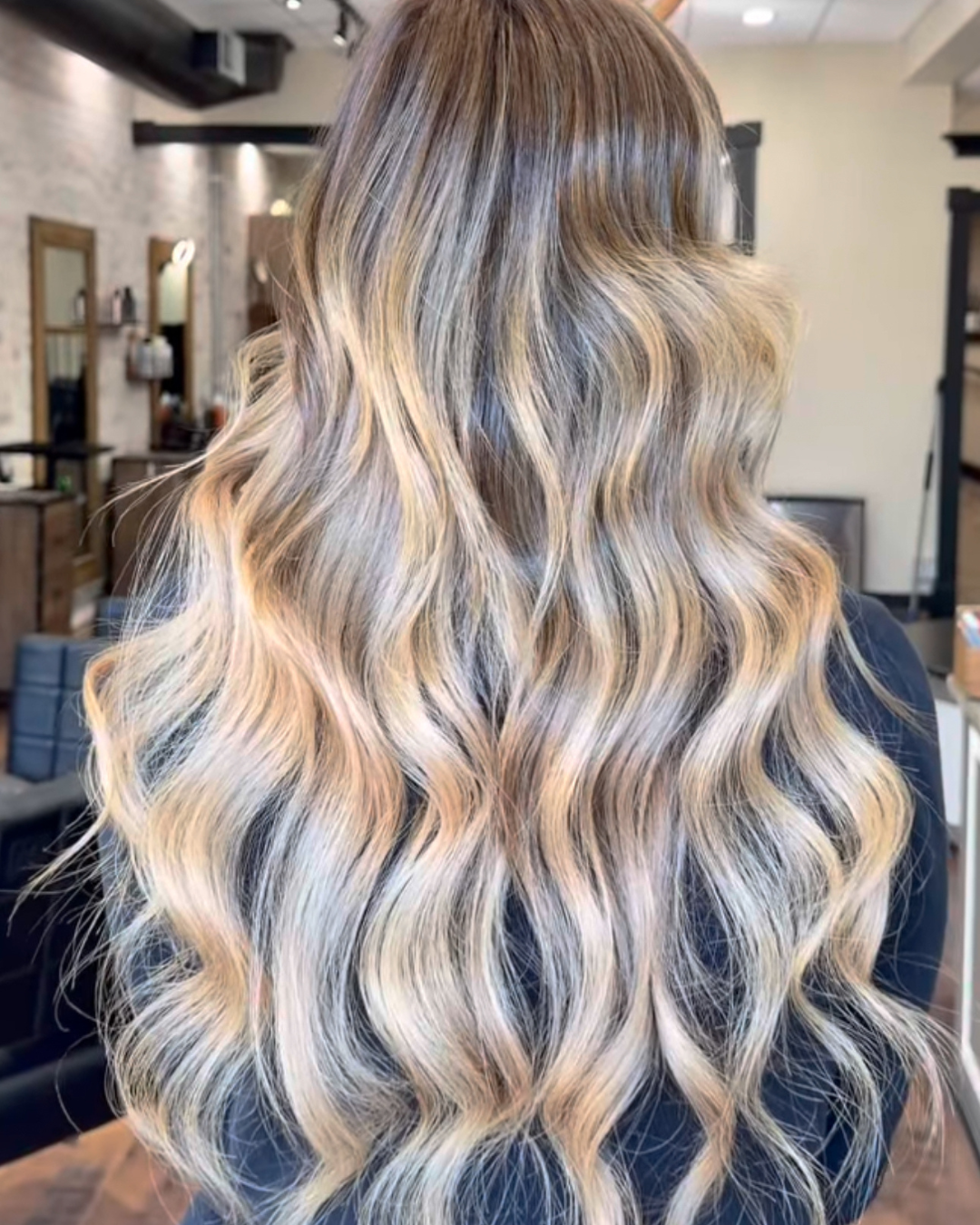 Back of woman's head showing long, wavy blonde hair with darker roots in a salon setting.