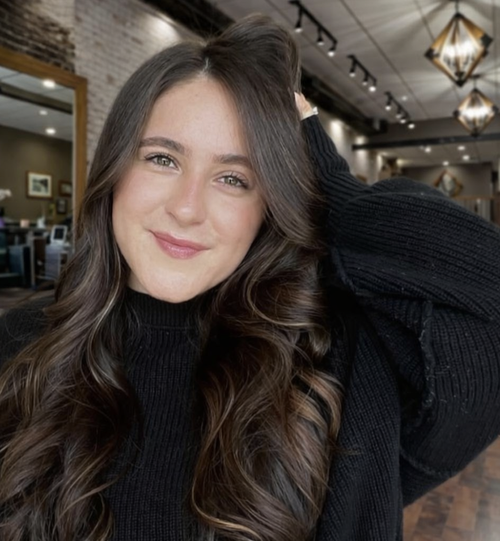 A young woman with long, wavy brown hair and a subtle smile, wearing a black sweater, seated in a warmly lit cafe or restaurant with modern decor.