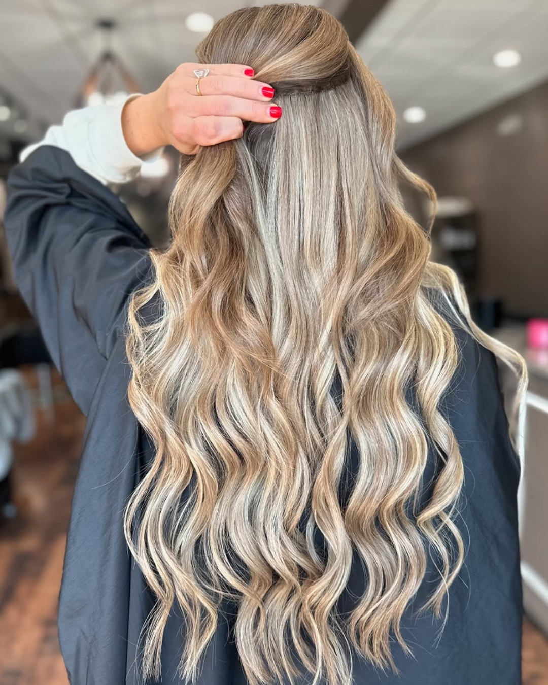 A woman with long, wavy, blonde hair and highlights touches her hair with one hand, showing red nail polish and a ring, in a salon or similar setting.
