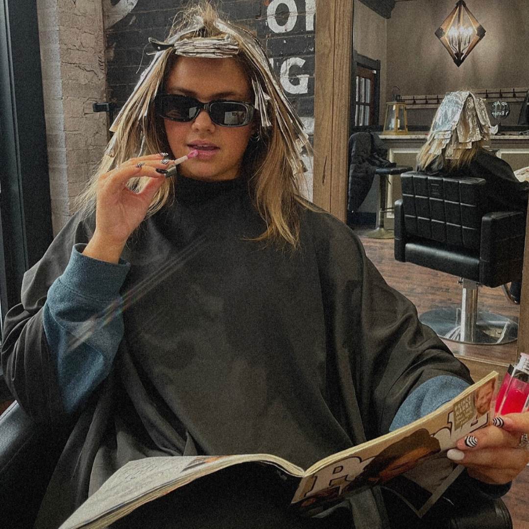 A woman with blonde hair and sunglasses sitting in a salon, holding a magazine in one hand and a cigarette in the other.