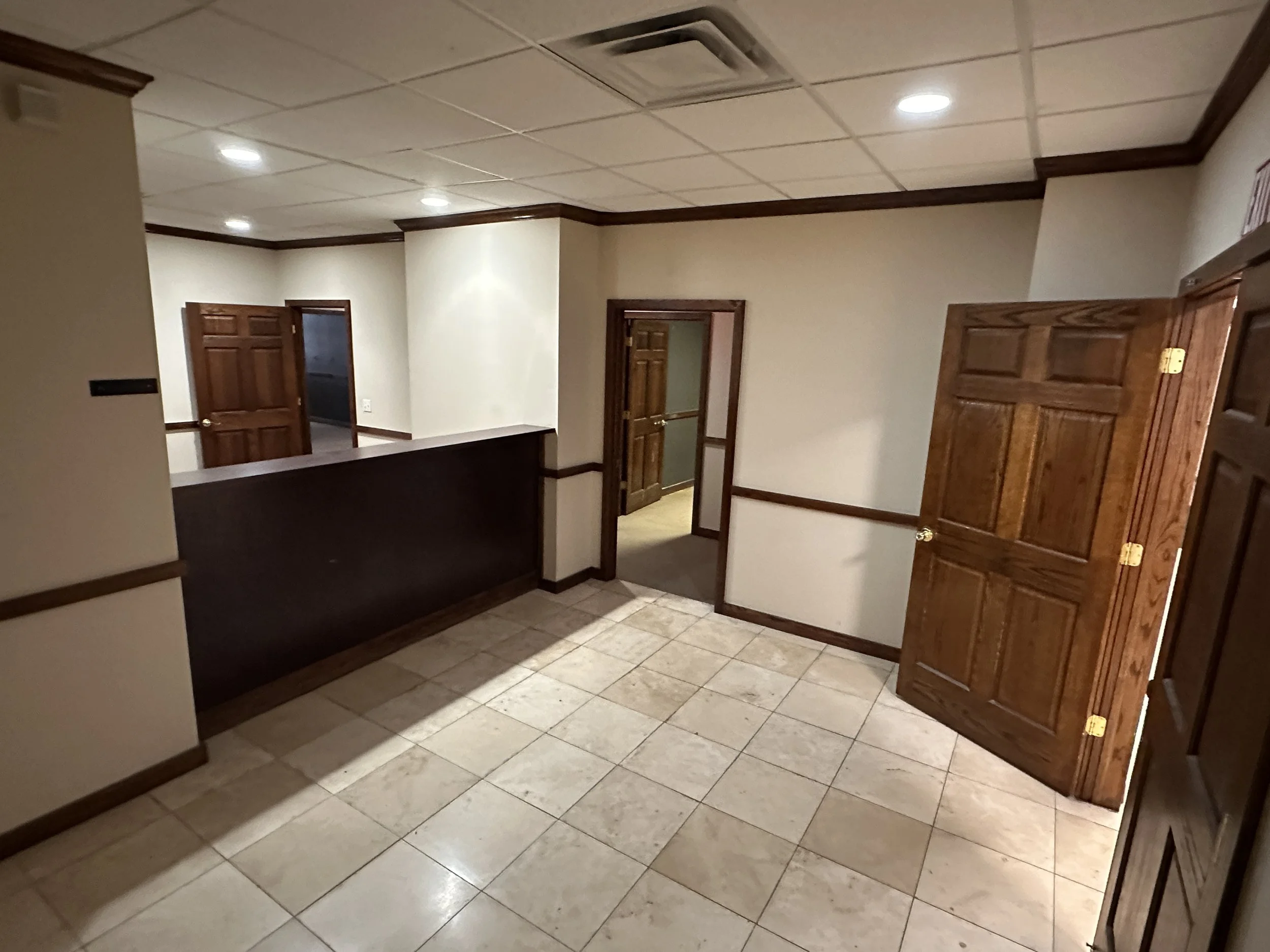 Empty office or lobby area with tiled flooring, beige walls with wooden trim, multiple wooden doors, and ceiling lights.