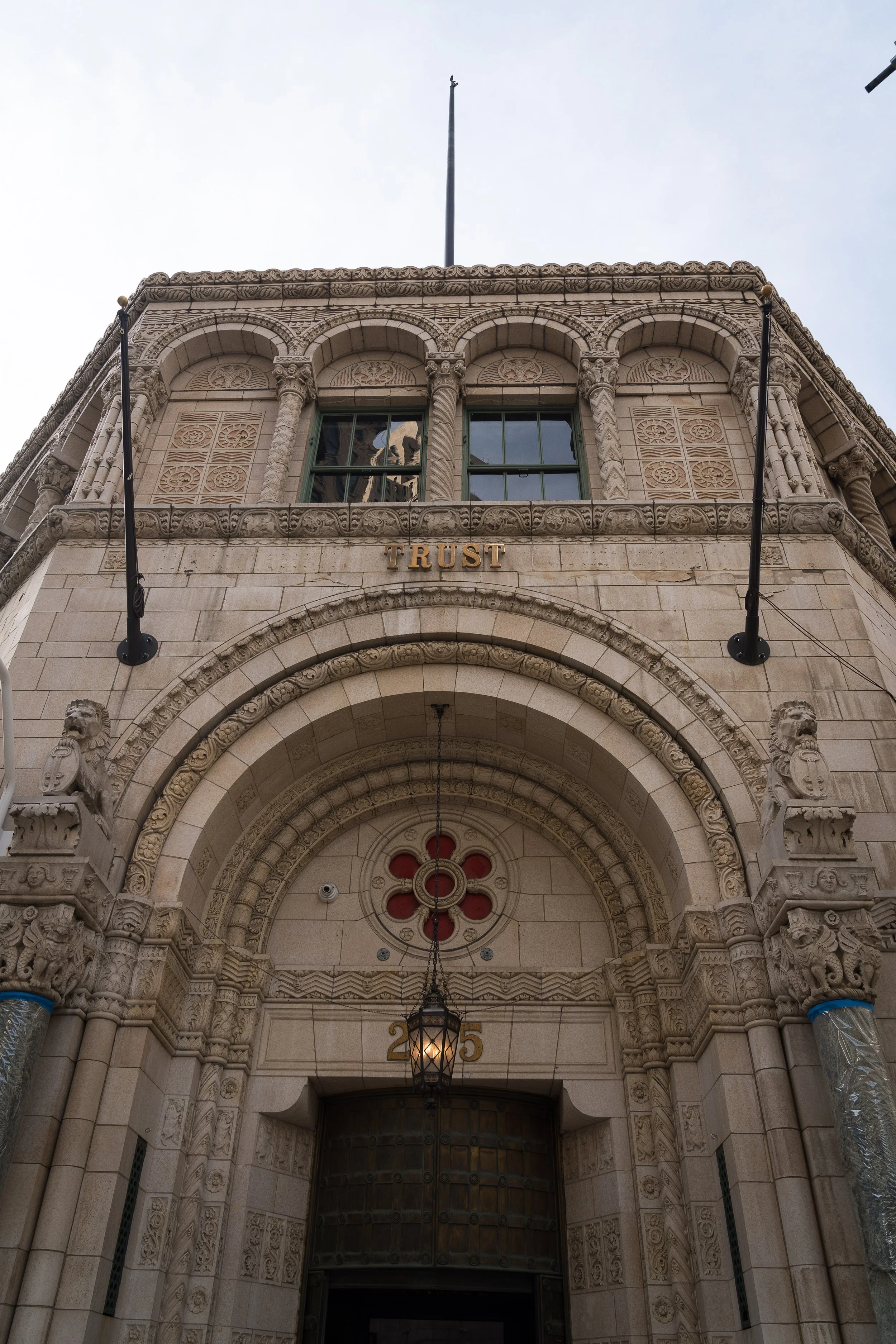 Front view of an ornate stone building with a sign that says 'TRUST' above the entrance, decorated with lions, a lamp hanging from the arch, and the number 205.