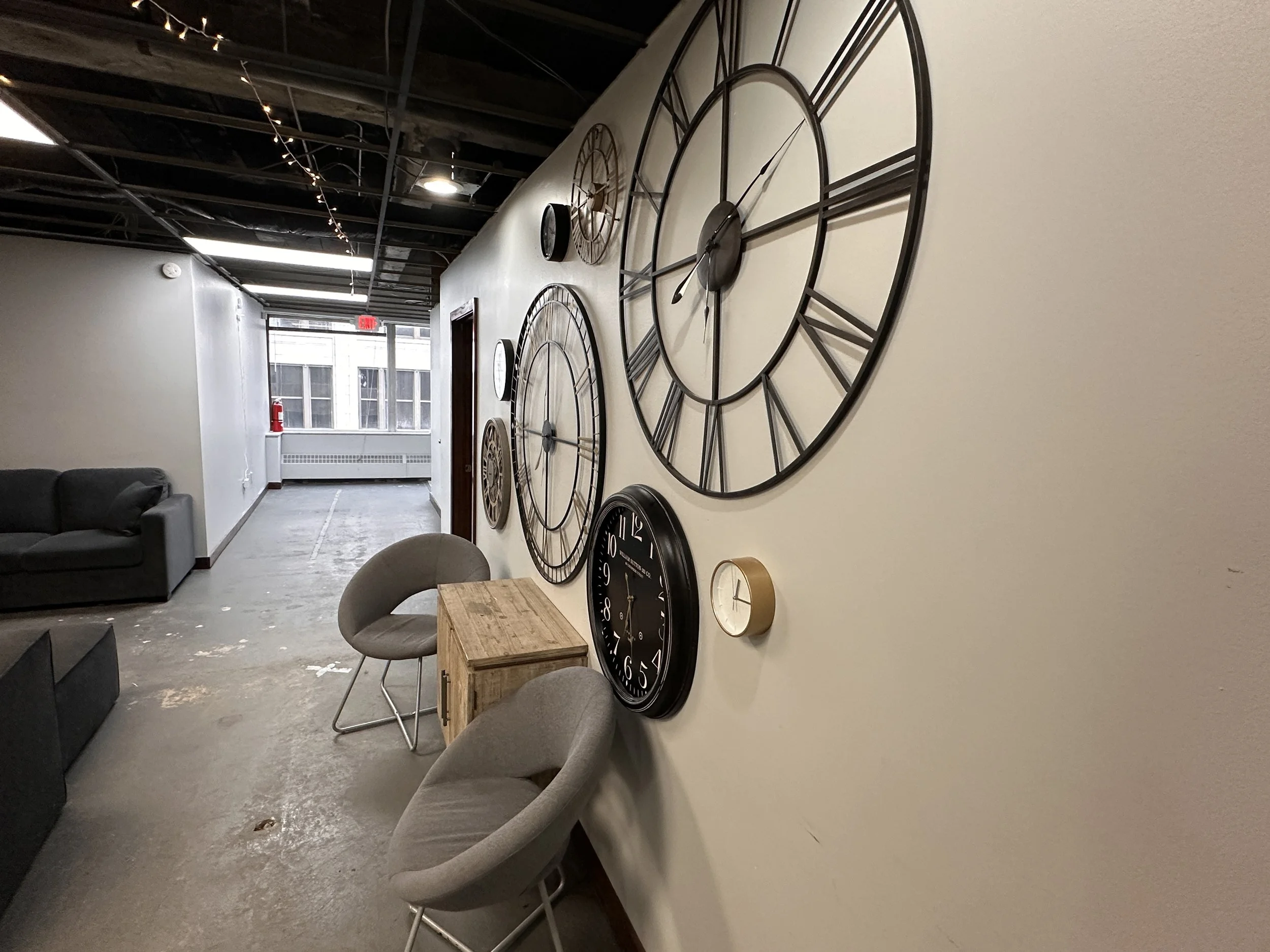 Interior of a modern lounge with a beige wall decorated with large black metal clocks; two gray upholstered chairs and a wooden side table in the foreground; exposed ceiling with string lights; hallway with a window at the end.