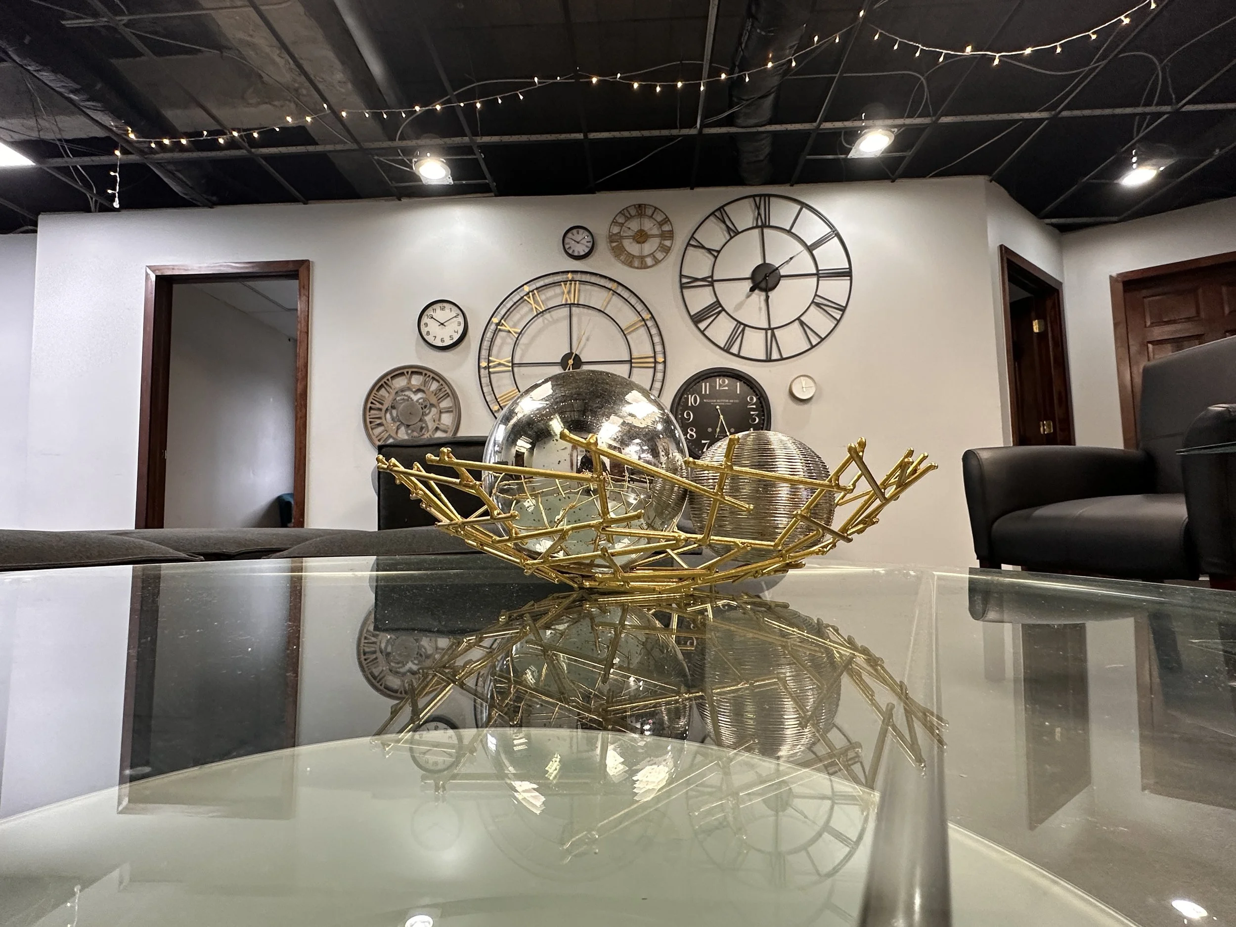 A room with a wall decorated with various clocks, a glass table with a decorative gold-colored bowl holding metallic spheres, and black chairs.