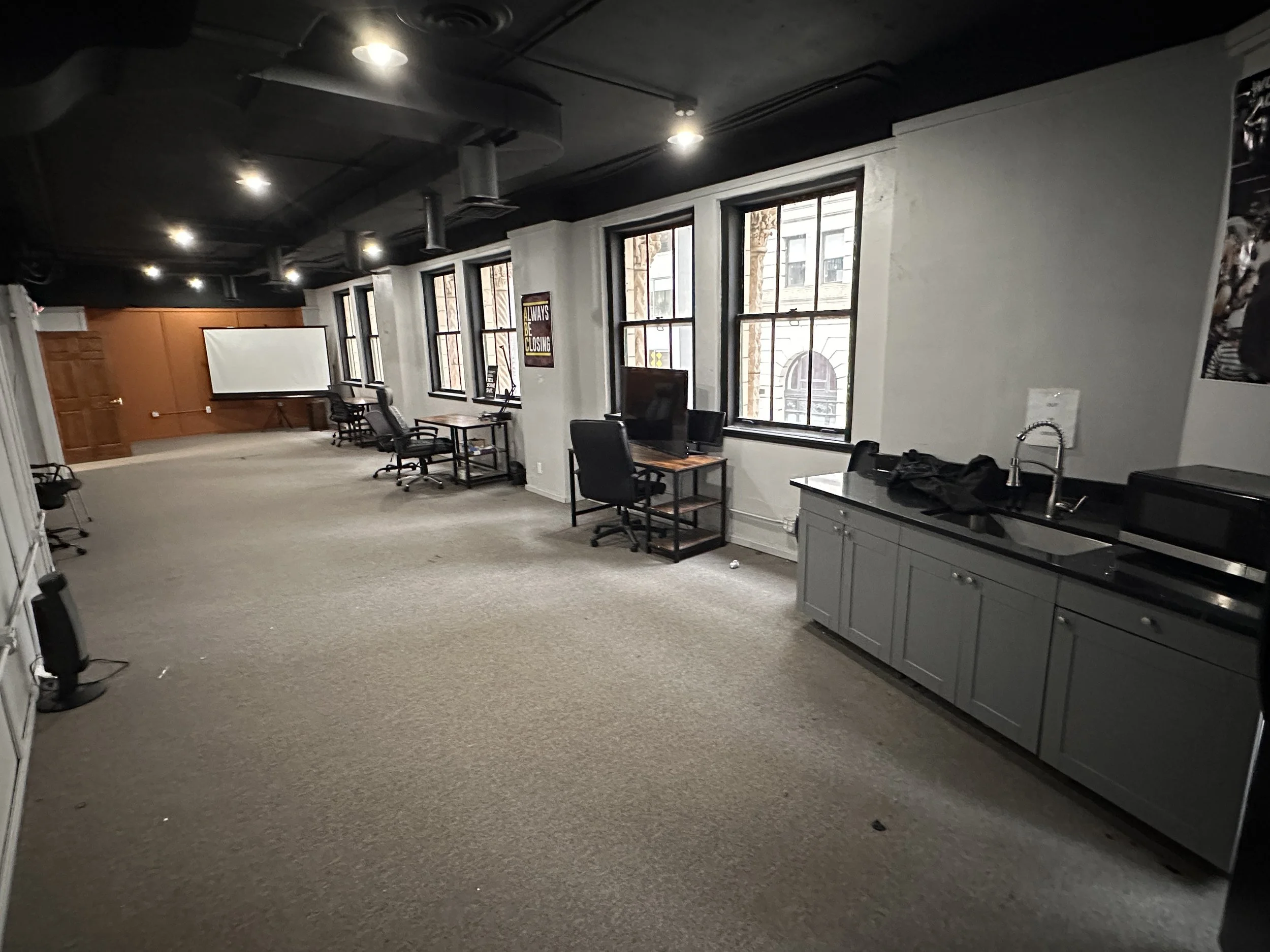 An empty office or conference room with several windows, chairs, desks, a whiteboard, and a kitchenette area with a sink and microwave.
