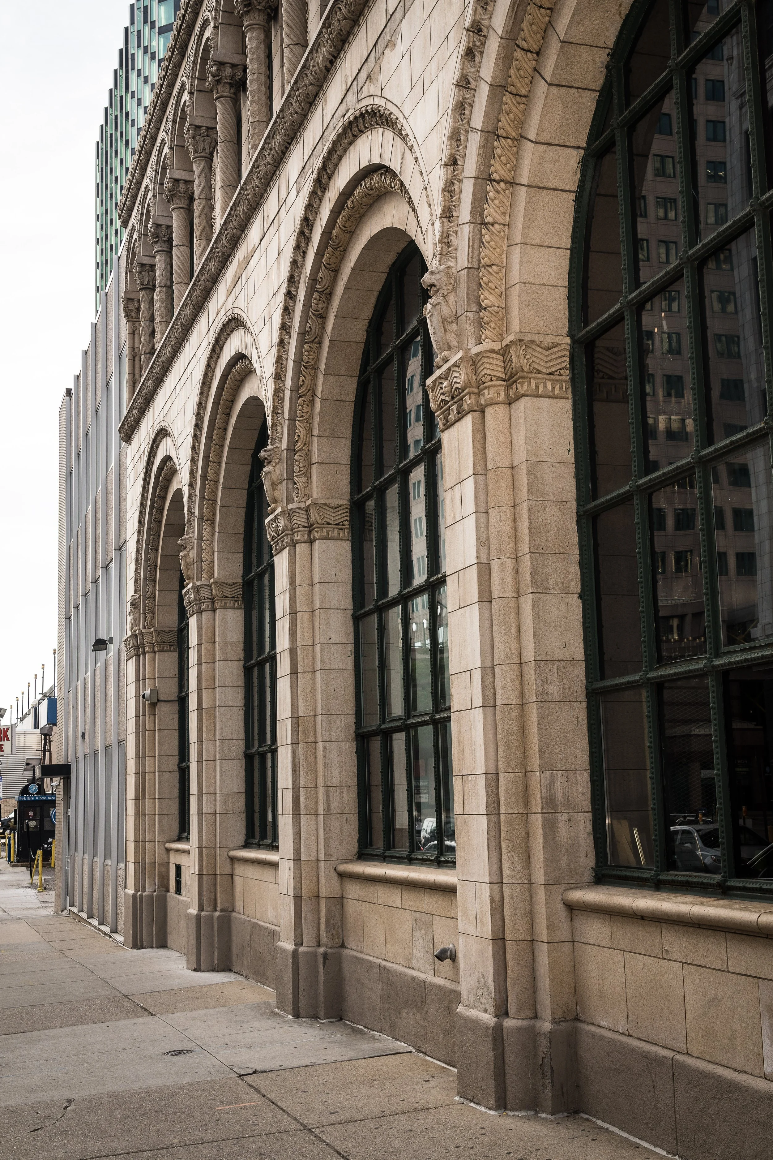 Exterior view of a historic building with stone arches, decorative columns, and large grid-pane windows on a city sidewalk.