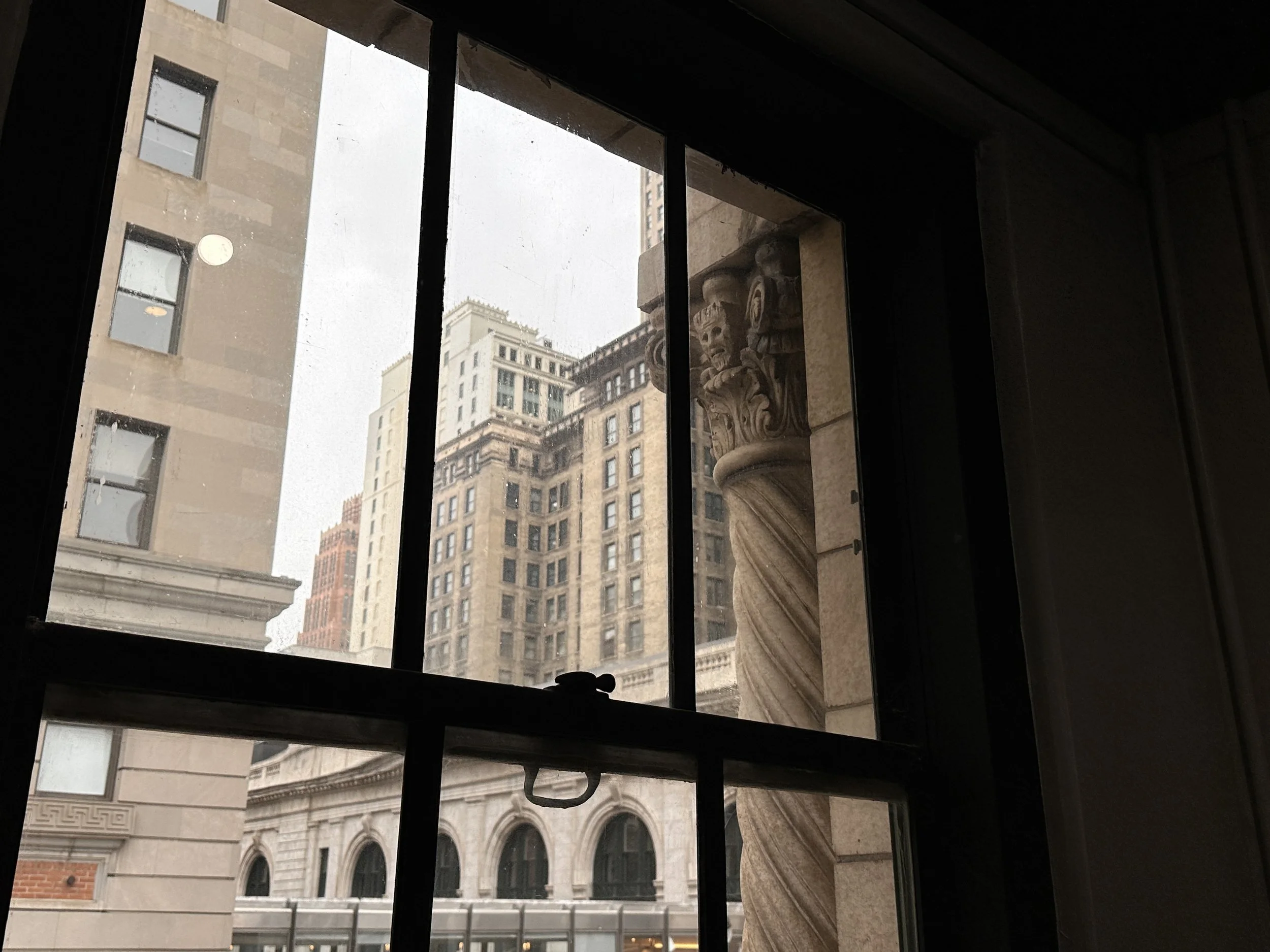 View of tall city buildings through a window with iron bars and decorative architectural column inside.