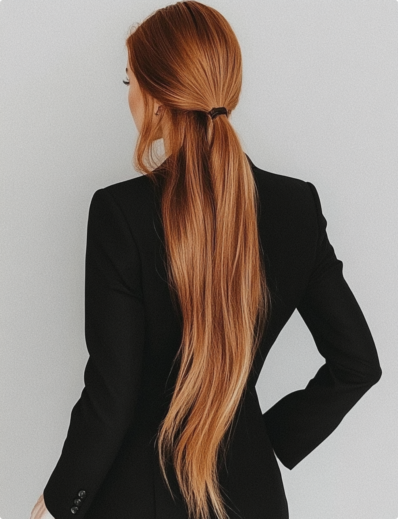 Back view of a woman with long, straight, red hair tied with a black hair tie, wearing a black blazer against a plain gray background.