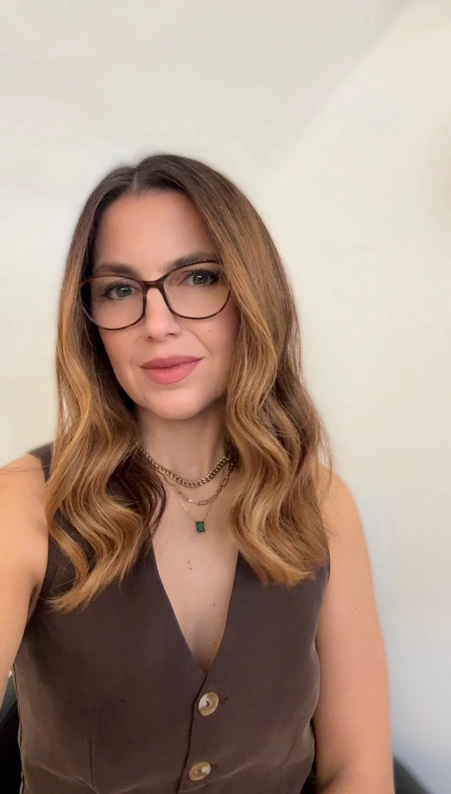 A woman with shoulder-length wavy light brown hair, wearing glasses, a brown sleeveless top with buttons, multiple necklaces including gold chains and a pendant, and natural makeup, standing against a plain white wall.