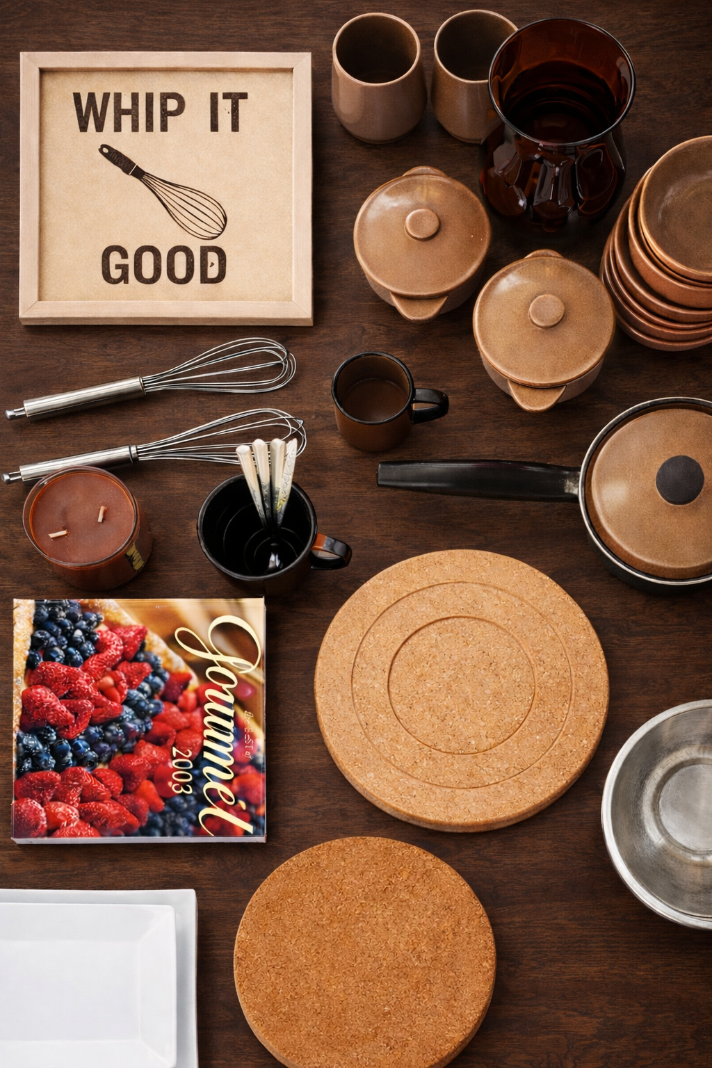 A kitchen countertop with pots, cups, plates, utensils, a cutting board, a cookbook, a candle, a sign reading 'Whip It Good' with a whisk, and a baking sheet.
