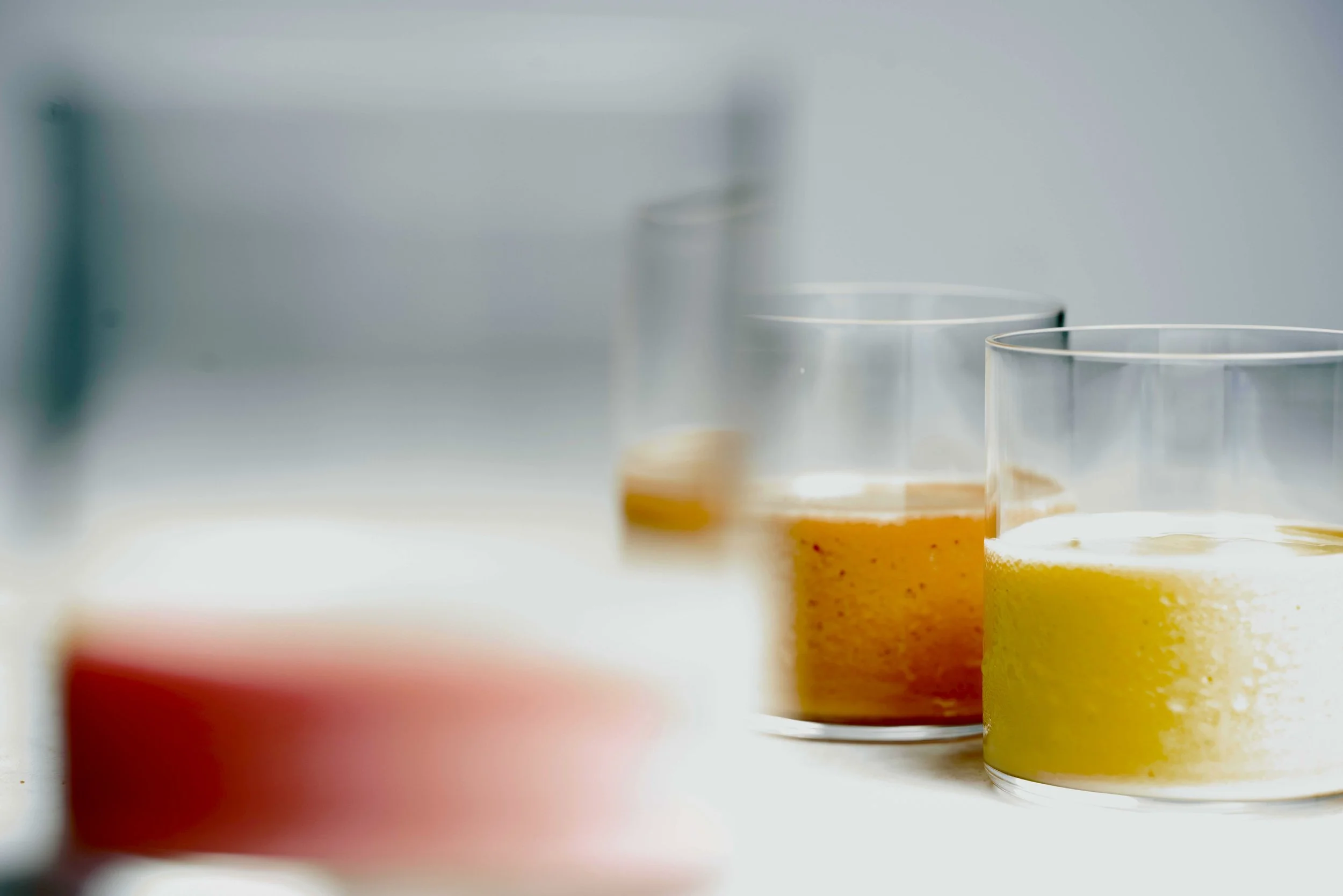Close-up of two glasses with yellow-orange liquid, possibly juice or a cocktail, on a white surface with a blurred background.