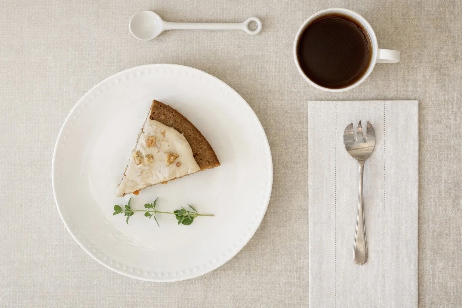 A slice of cheesecake on a white plate garnished with a sprig of herbs, placed on a beige table. Next to the plate is a cup of black coffee, a white napkin with a fork on top, and a small white spoon.