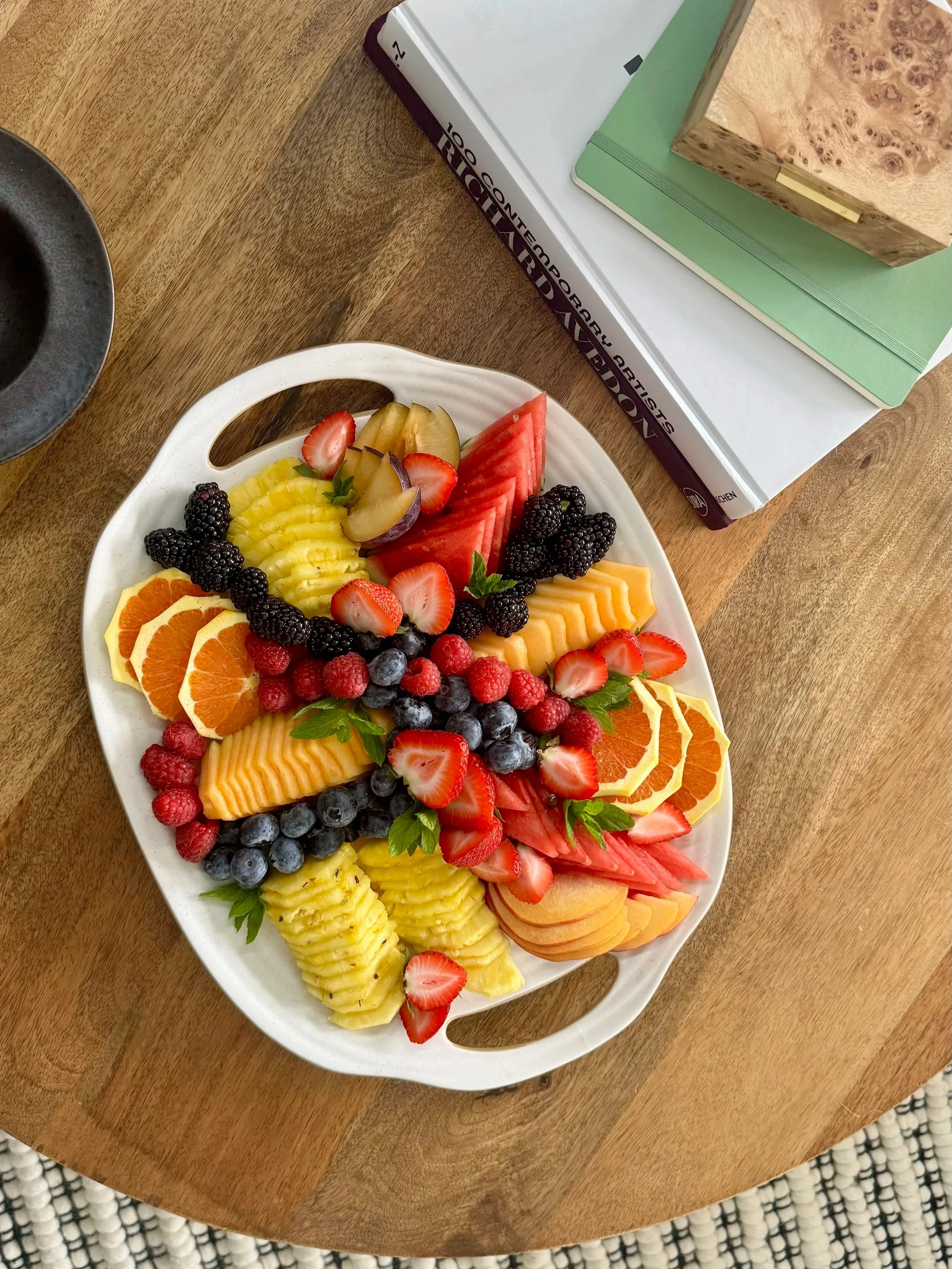 A white plate filled with a variety of fresh sliced fruits including oranges, strawberries, blueberries, raspberries, blackberries, pineapple, watermelon, honeydew, cantaloupe, and pear. The plate sits on a wooden table.