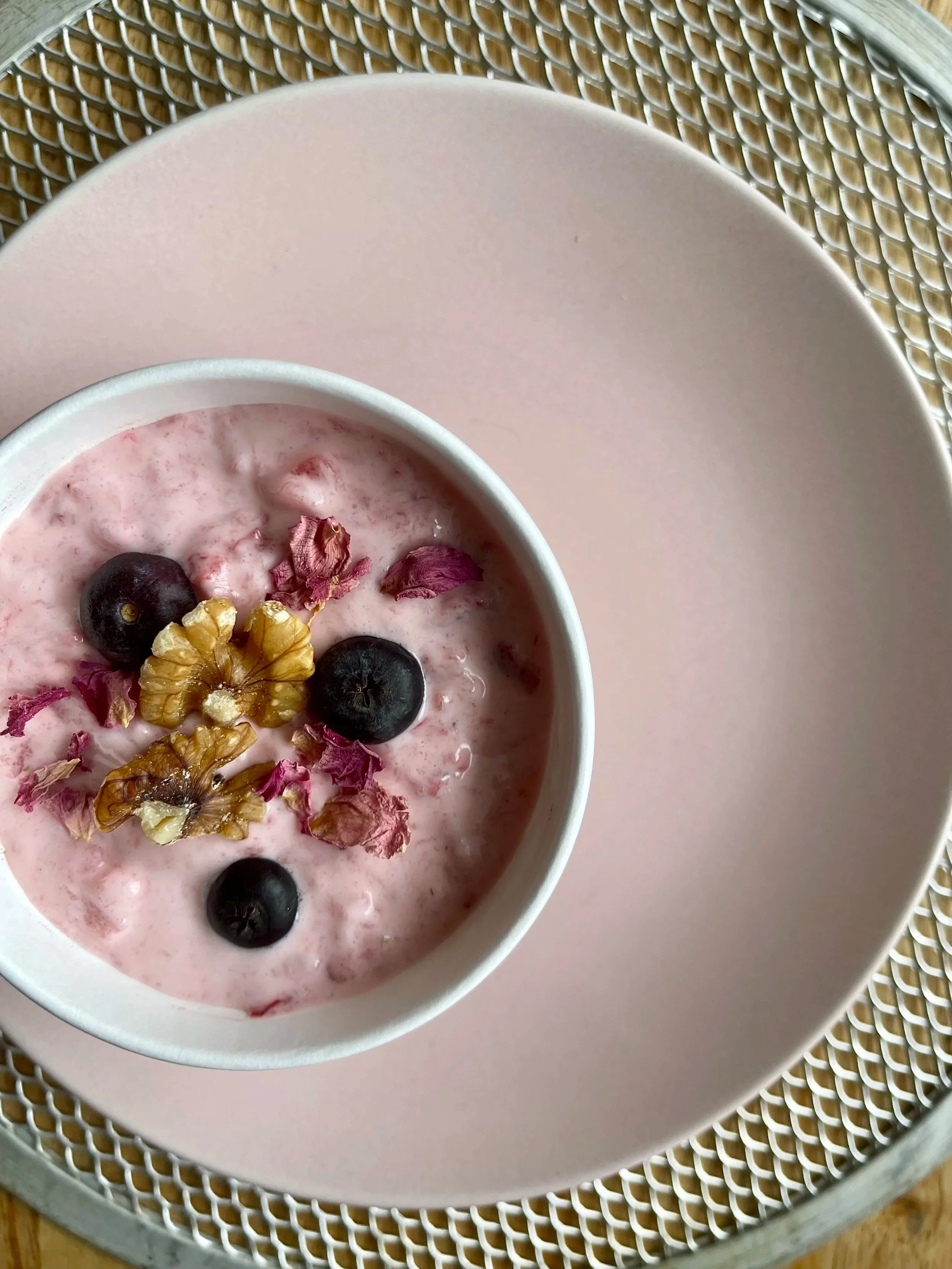 A bowl of pink yogurt topped with blueberries, walnuts, and dried rose petals, sitting on a pink plate on a metal table with a woven pattern.
