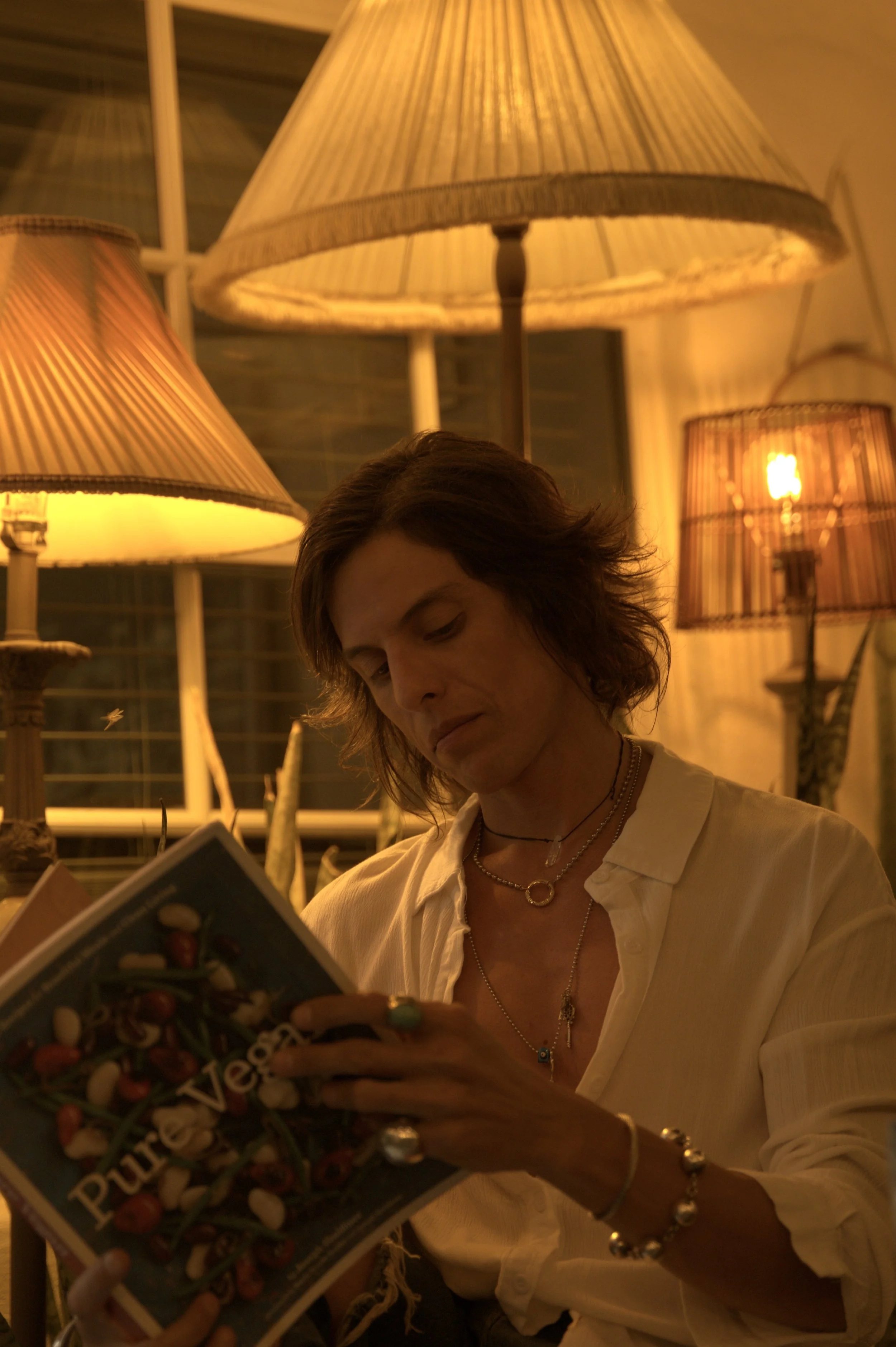 A woman with wavy hair reads a book under warm yellow-orange lighting from multiple lamps in a cozy indoor setting.