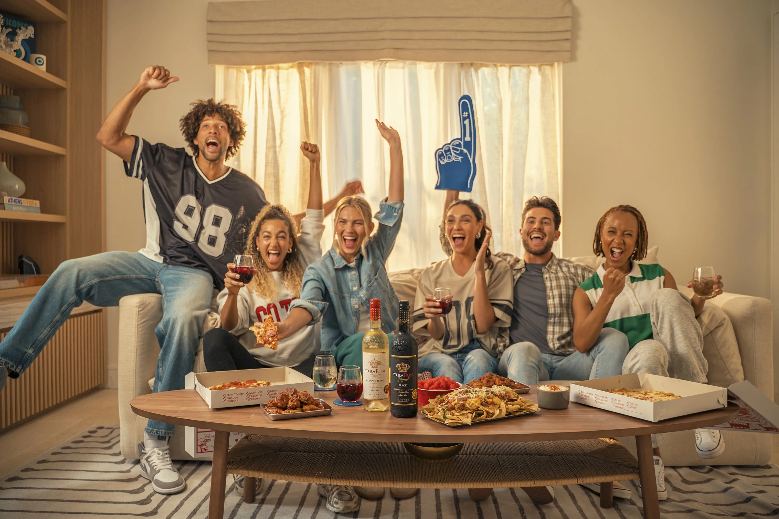 A group of six friends sitting on a couch celebrating with pizza, wine, and snacks in a living room, with some raising their arms and laughing.