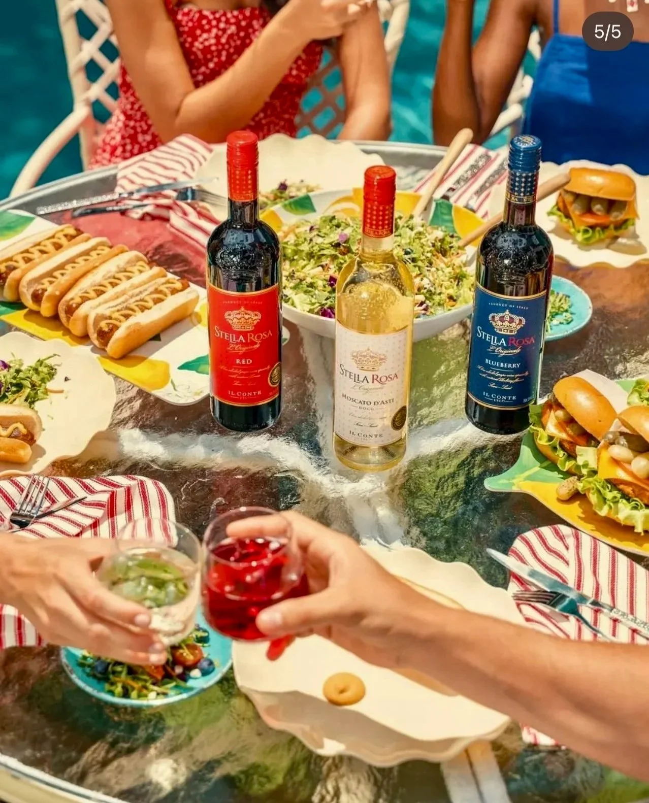 A table set with various foods and drinks at an outdoor gathering. There are three bottles of Stella Rosa wine, a salad, mini sandwiches, and small burger sliders. People are reaching for drinks, with one holding a glass of red beverage and another with a wine glass. The scene suggests a casual summer meal or party.