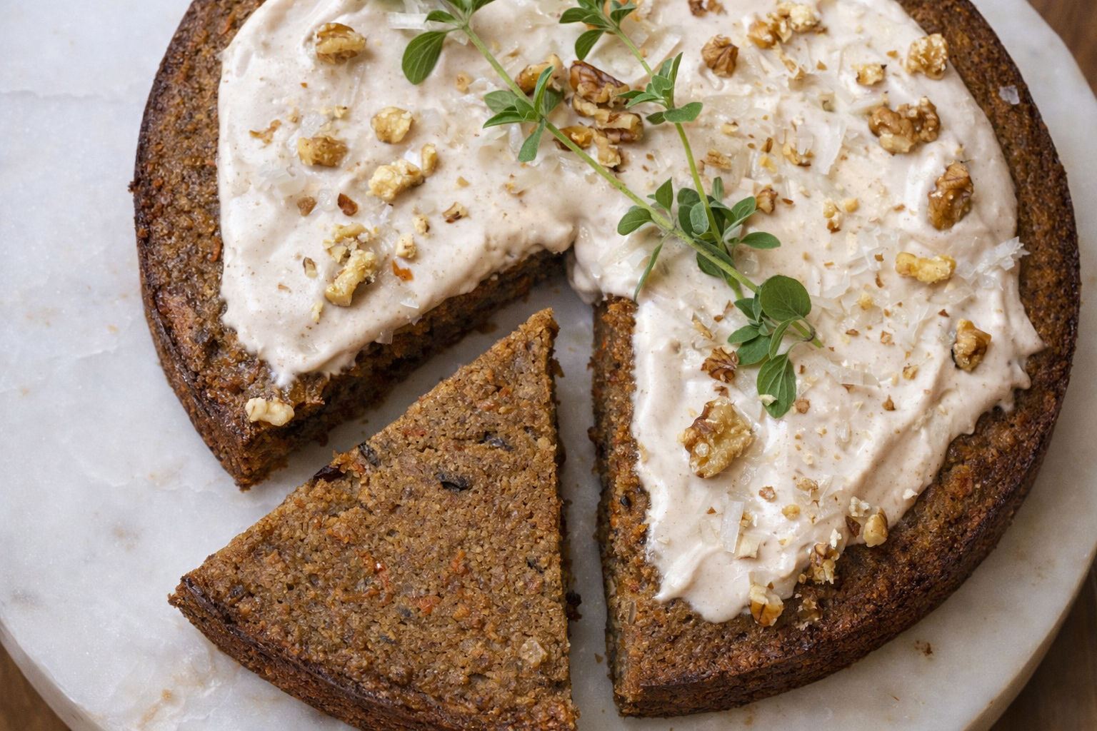 A sliced carrot cake with cream cheese frosting, topped with chopped walnuts and garnished with fresh thyme on a marble surface.