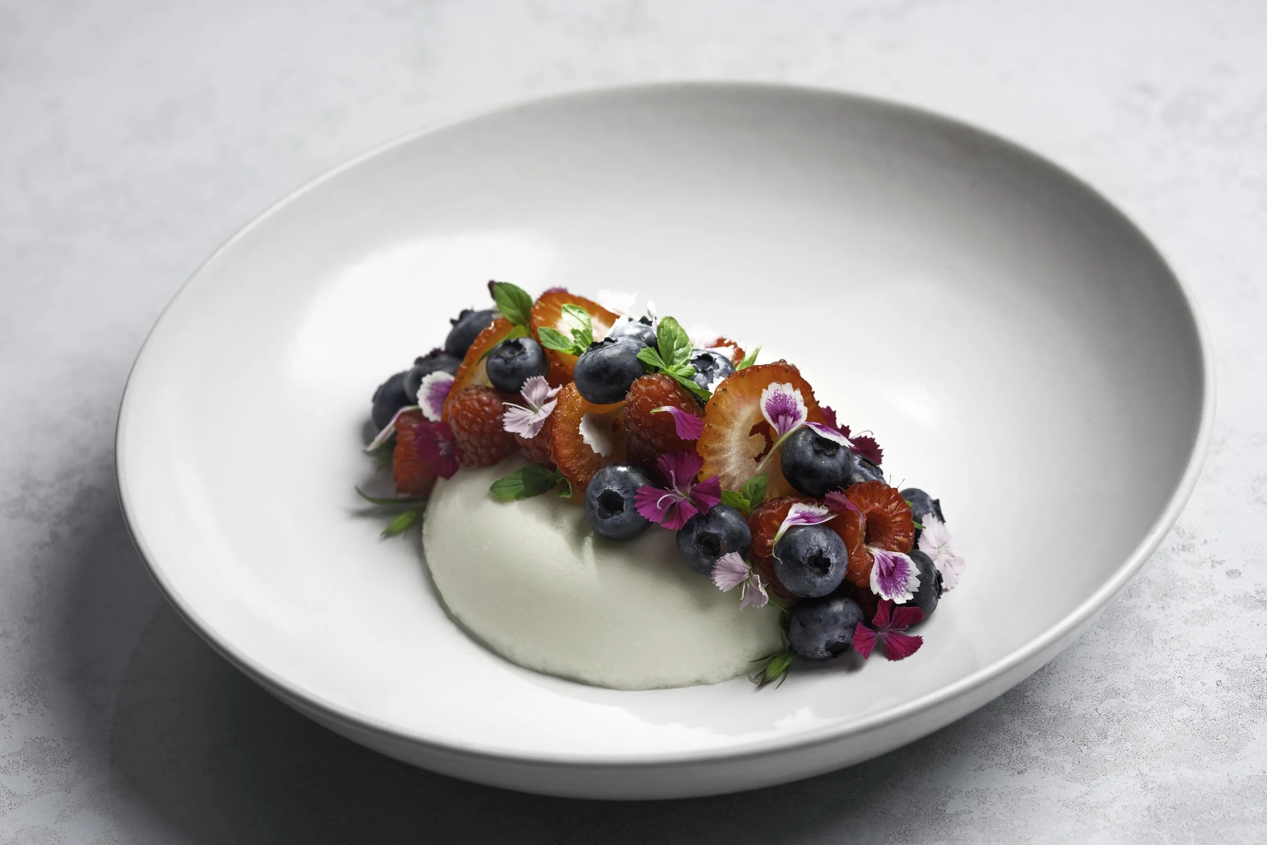 Dessert with mixed berries, edible flowers, and fresh herbs on a white plate