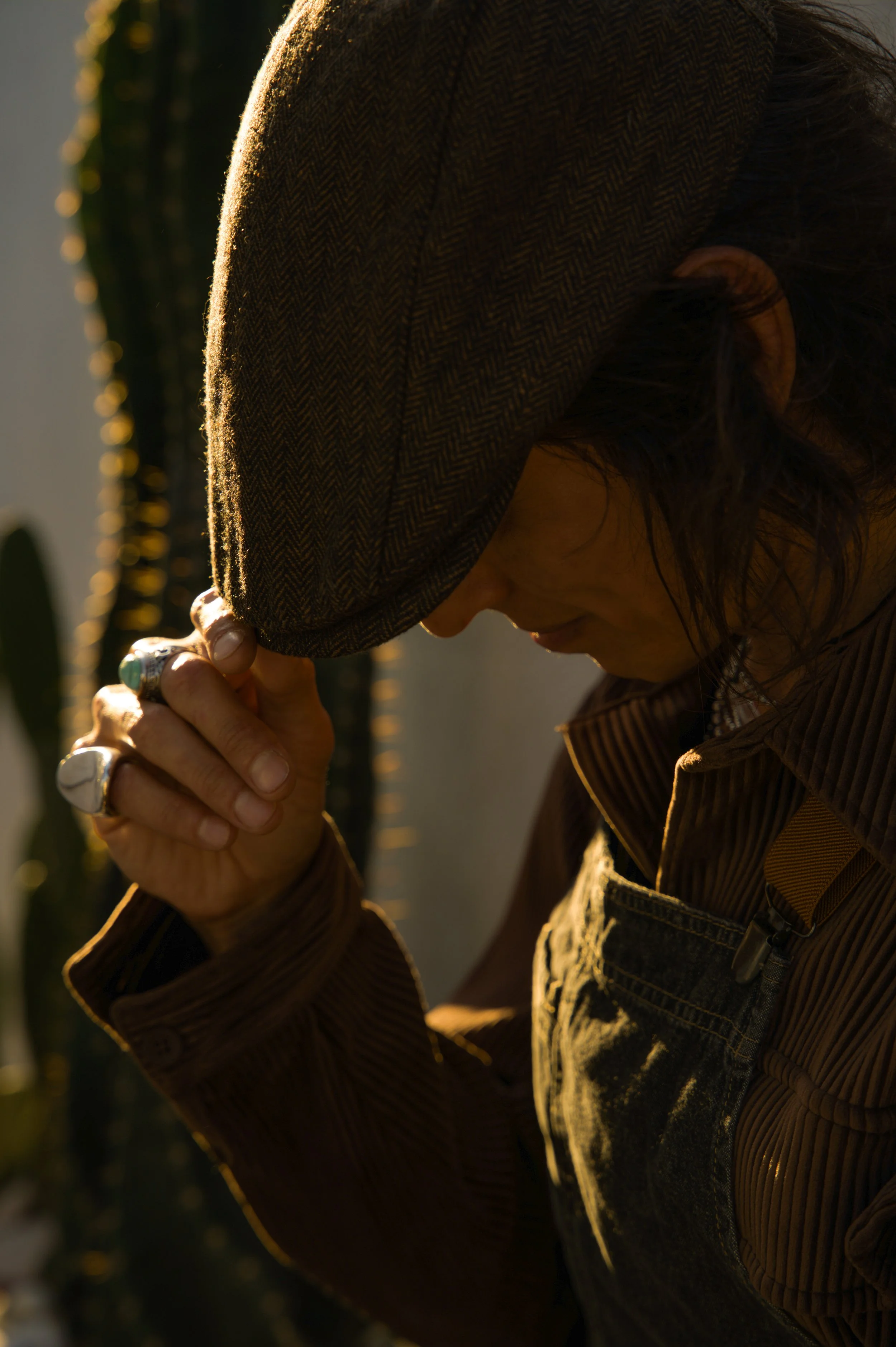 A person wearing a brown herringbone hat and brown striped shirt, with dark curly hair, bowing their head and clasping their hands together in prayer or reflection, illuminated by warm sunlight.