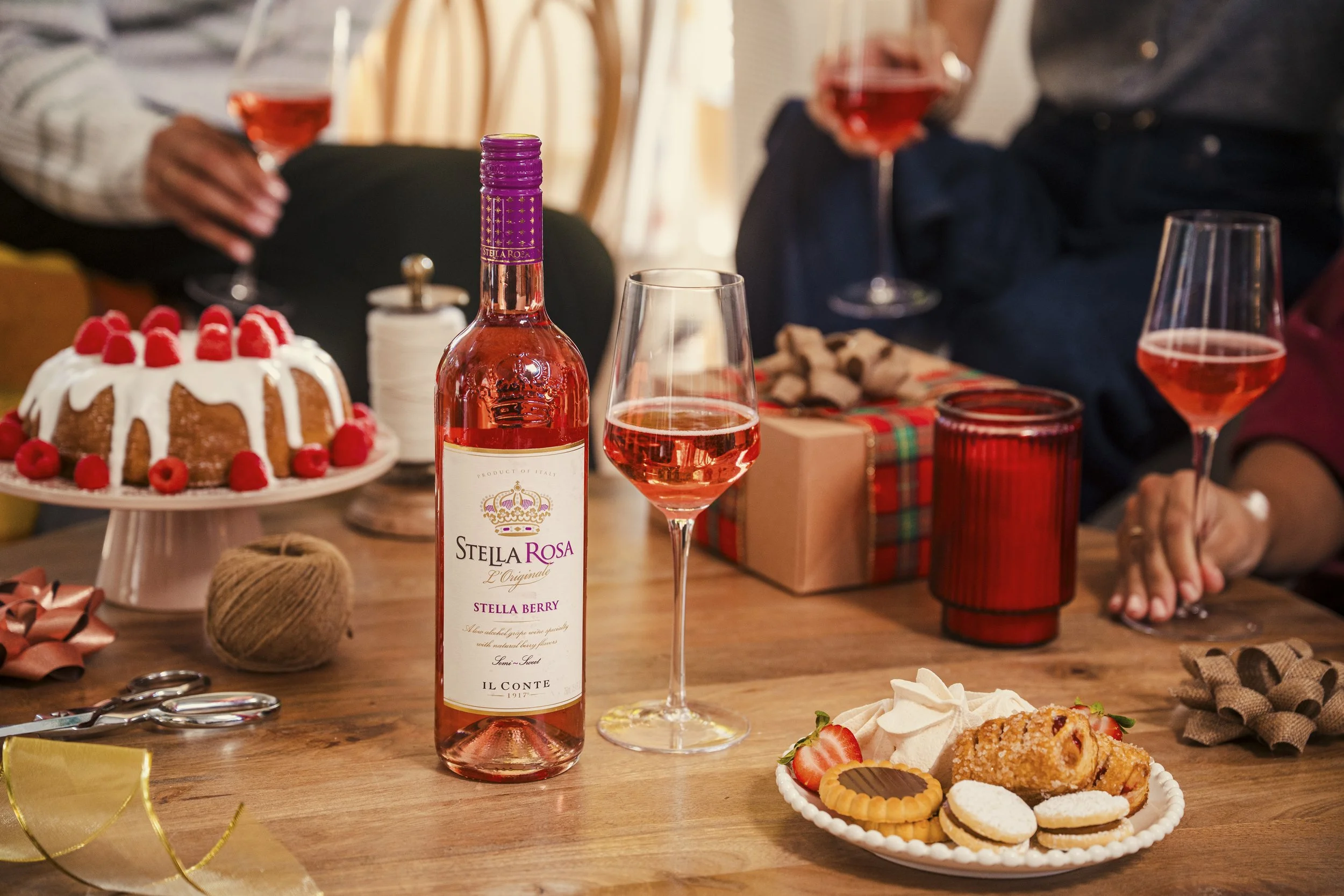 A table with a bottle of Stella Rosa wine, a glass of rosé wine, a decorated cake with strawberries and raspberries, assorted cookies and strawberries on a small plate, and holiday decorations in the background among people holding glasses of rosé wine.