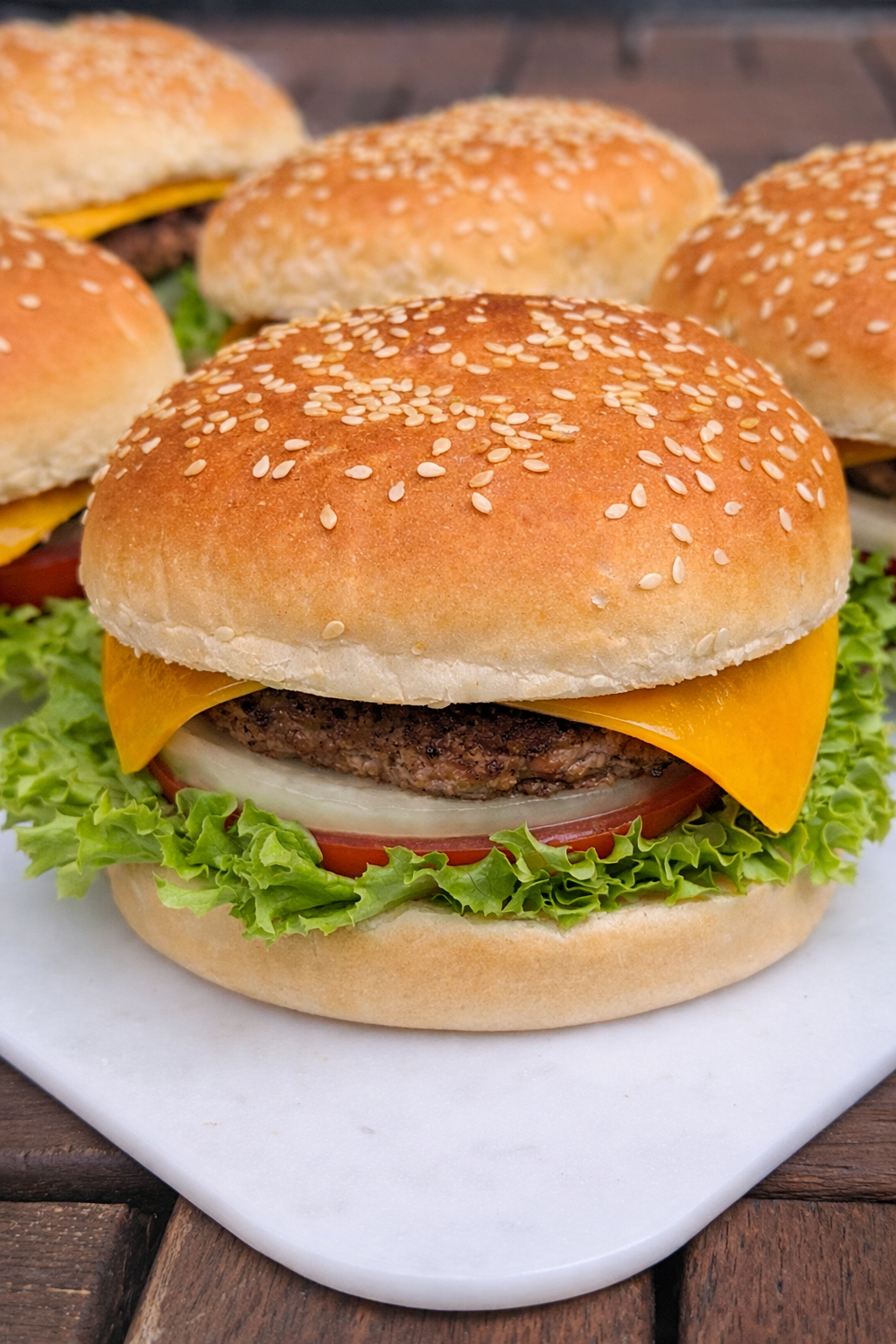 Close-up of a cheeseburger with lettuce, tomato, onion, a beef patty, melted cheddar cheese, and a sesame seed bun on a white plate.