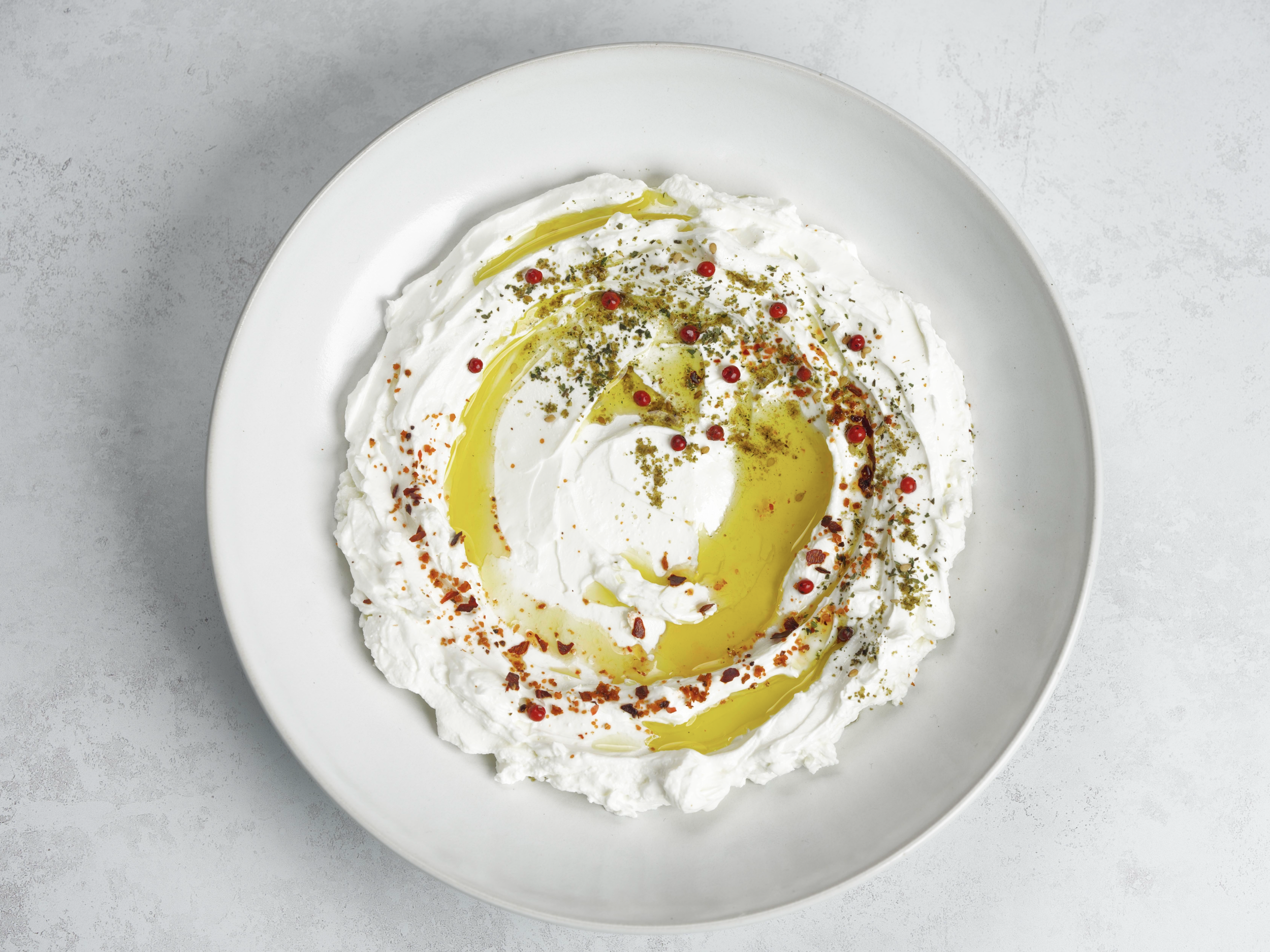A white plate with a baked dip topped with olive oil, red pepper flakes, black pepper, herbs, and small red peppercorns, on a gray surface.
