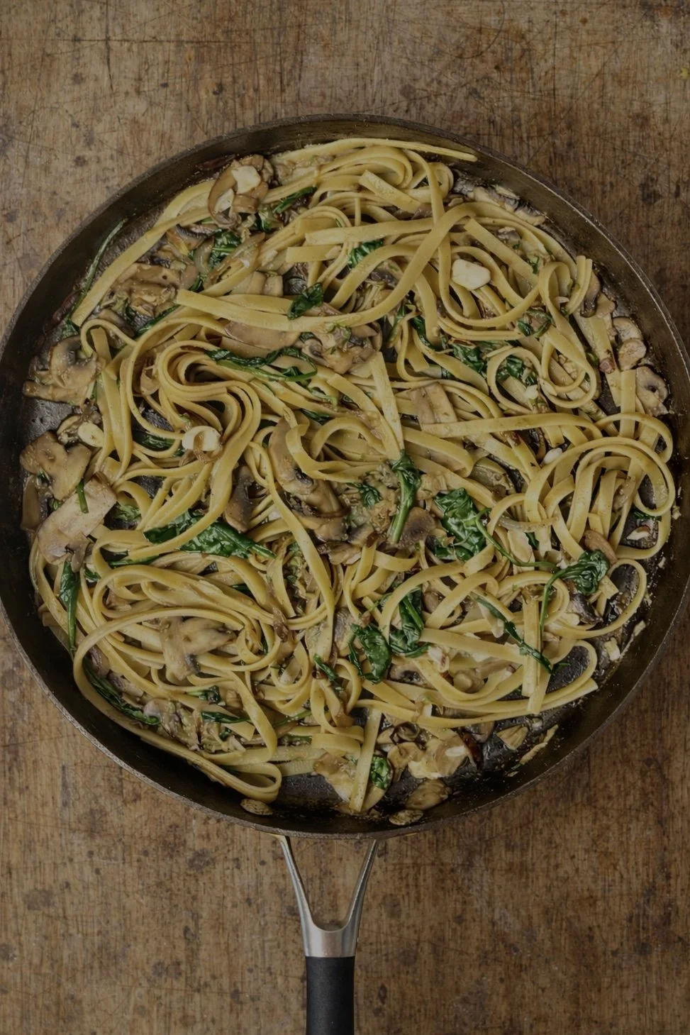 Fettuccine pasta with mushrooms, spinach, and garlic in a skillet on a wooden surface.