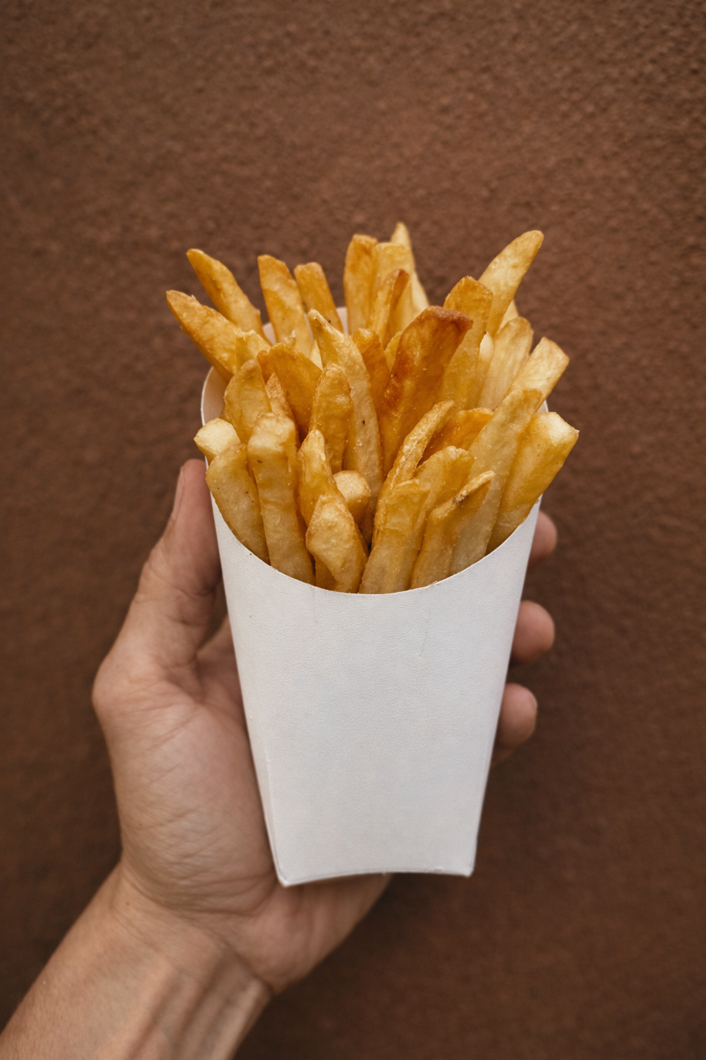 A hand holding a white paper cone of French fries against a brown background.