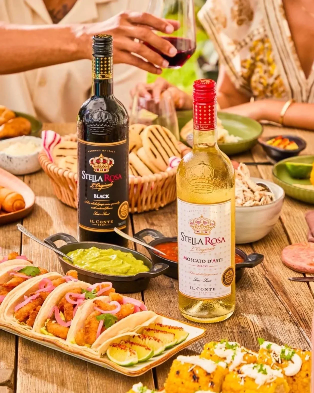 People enjoying a meal with various dishes including tacos, guacamole, and sauces, along with bottles of Stella Rosa wine on a wooden table.