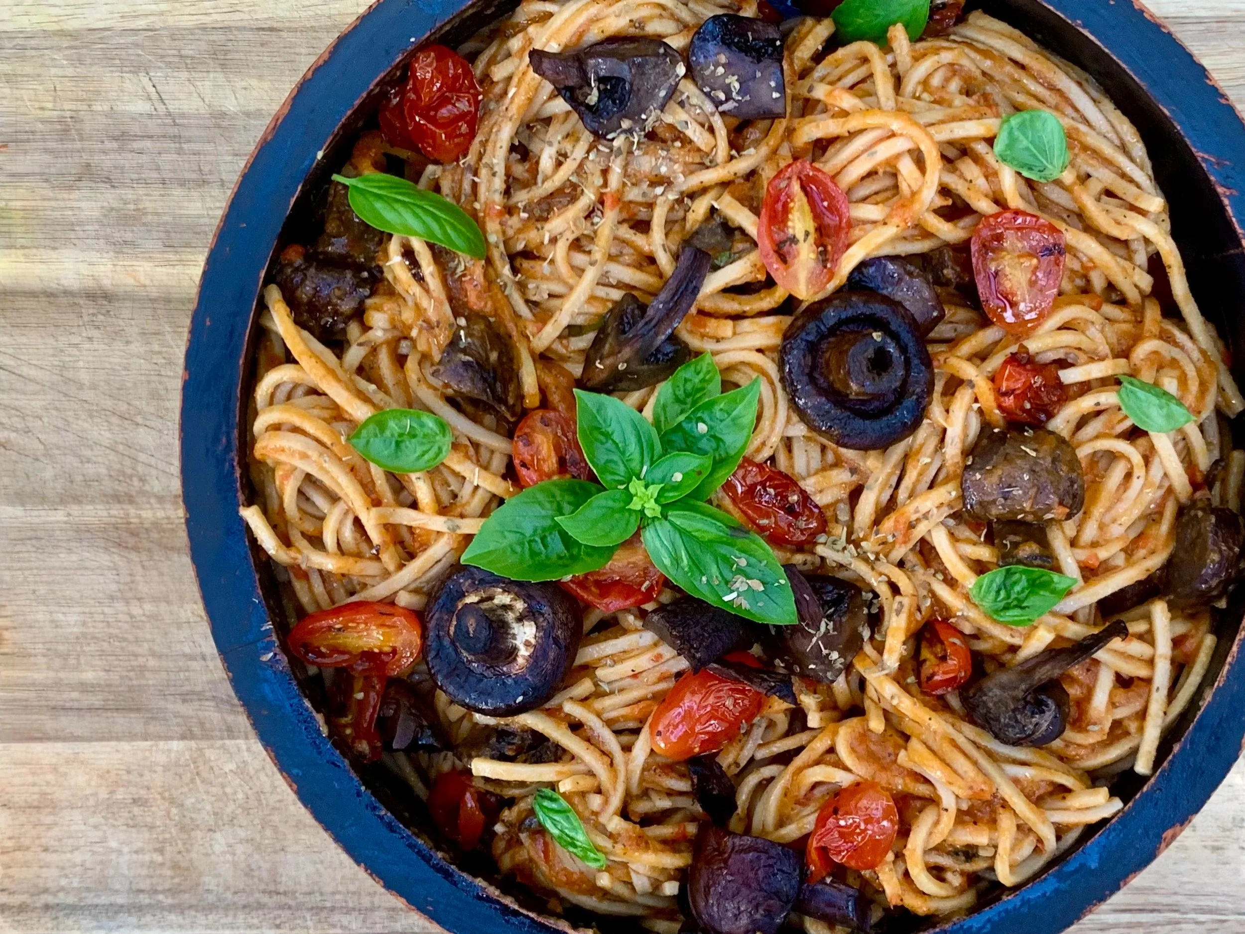 A bowl of spaghetti pasta with cherry tomatoes, black olives, basil leaves, and grated cheese, placed on a wooden surface.