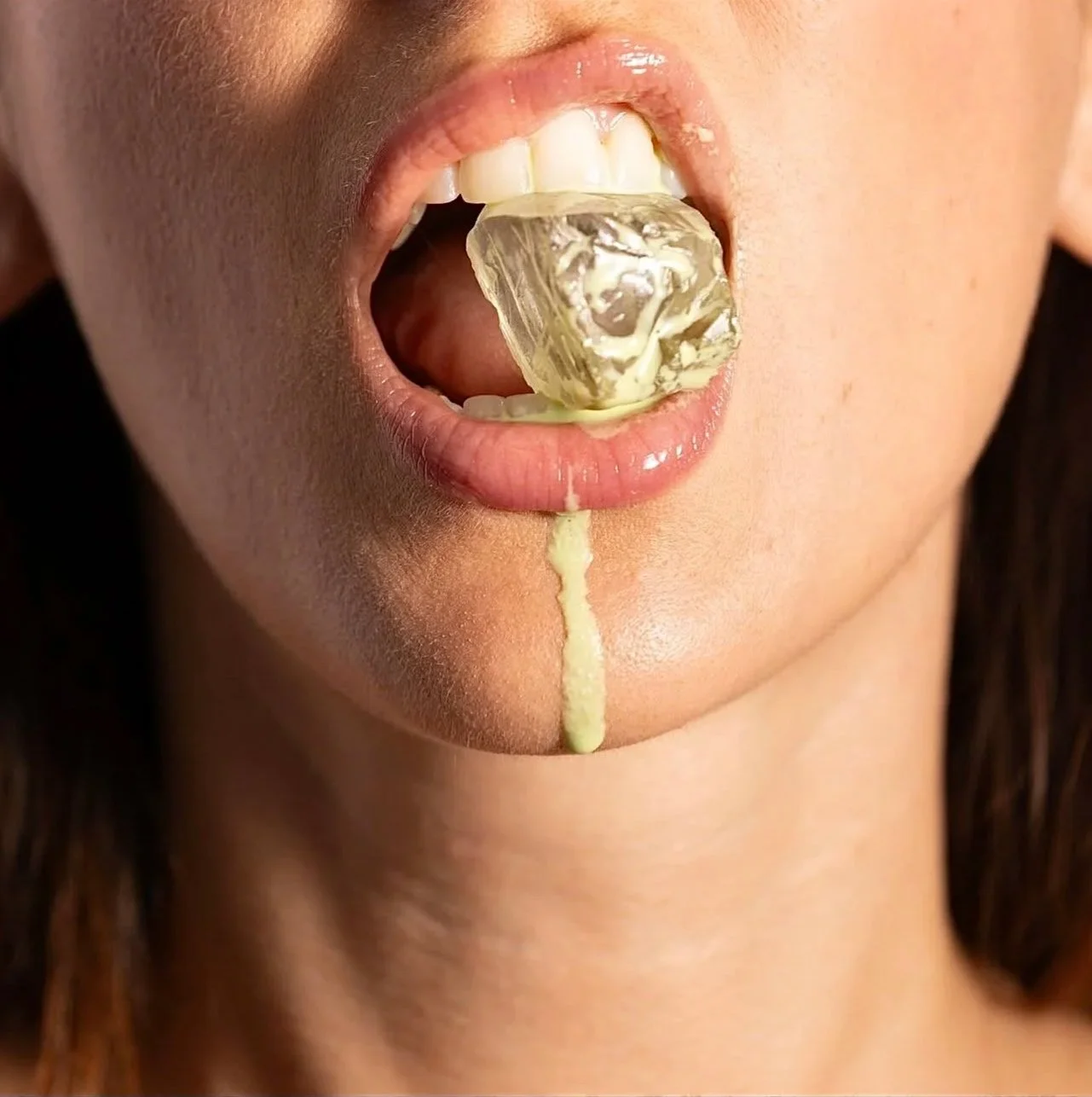 Close-up of a woman's face with her mouth open, holding a square ice cube on her tongue with melting yellowish substance dripping down her chin.