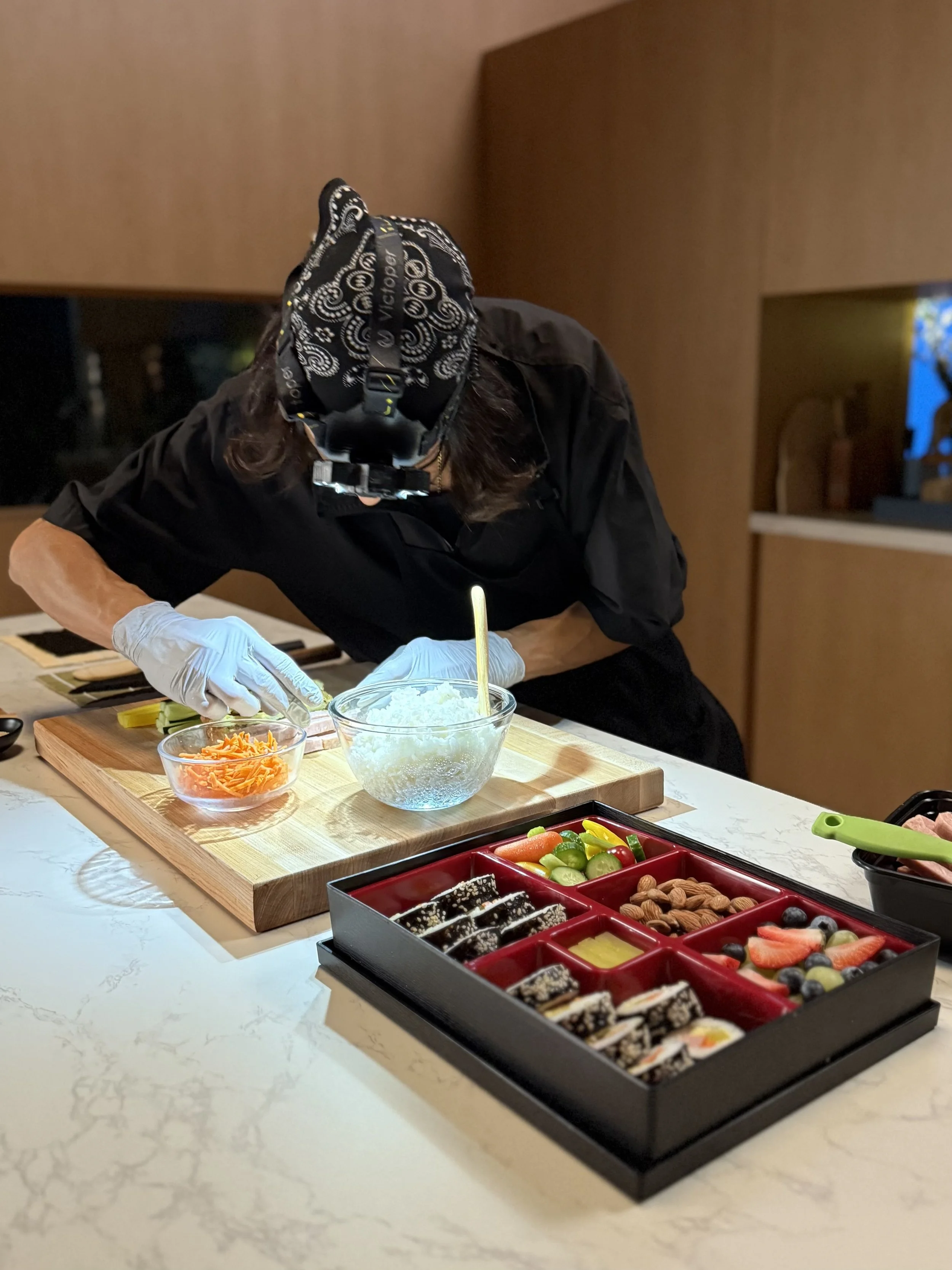 Person preparing sushi ingredients on a wooden cutting board, with bowls of shredded carrots, rice, and a tray of assorted sushi and fruits on a marble countertop.