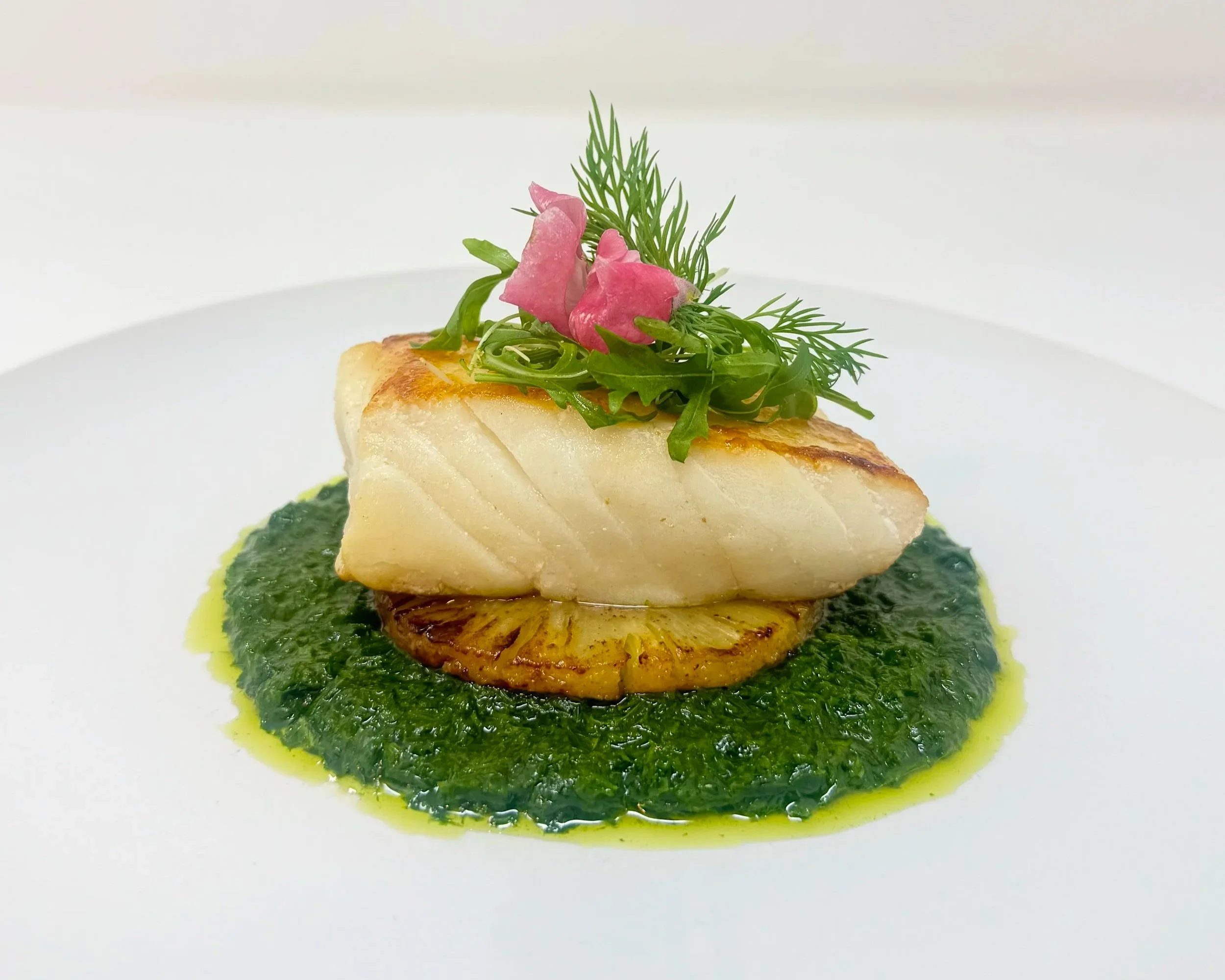A gourmet dish featuring a white fish fillet topped with pink pickled onions and fresh herbs, served on a bed of green sauce on a white plate.