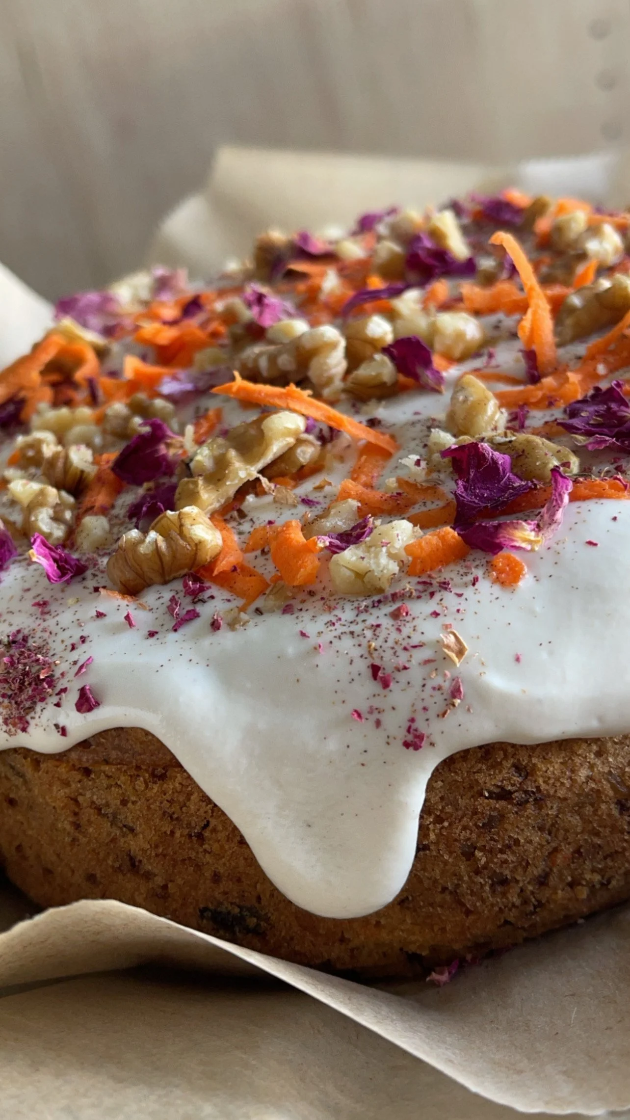 Close-up of a cake with white frosting, topped with chopped walnuts, shredded carrots, purple flower petals, and pinkish-red sprinkles.