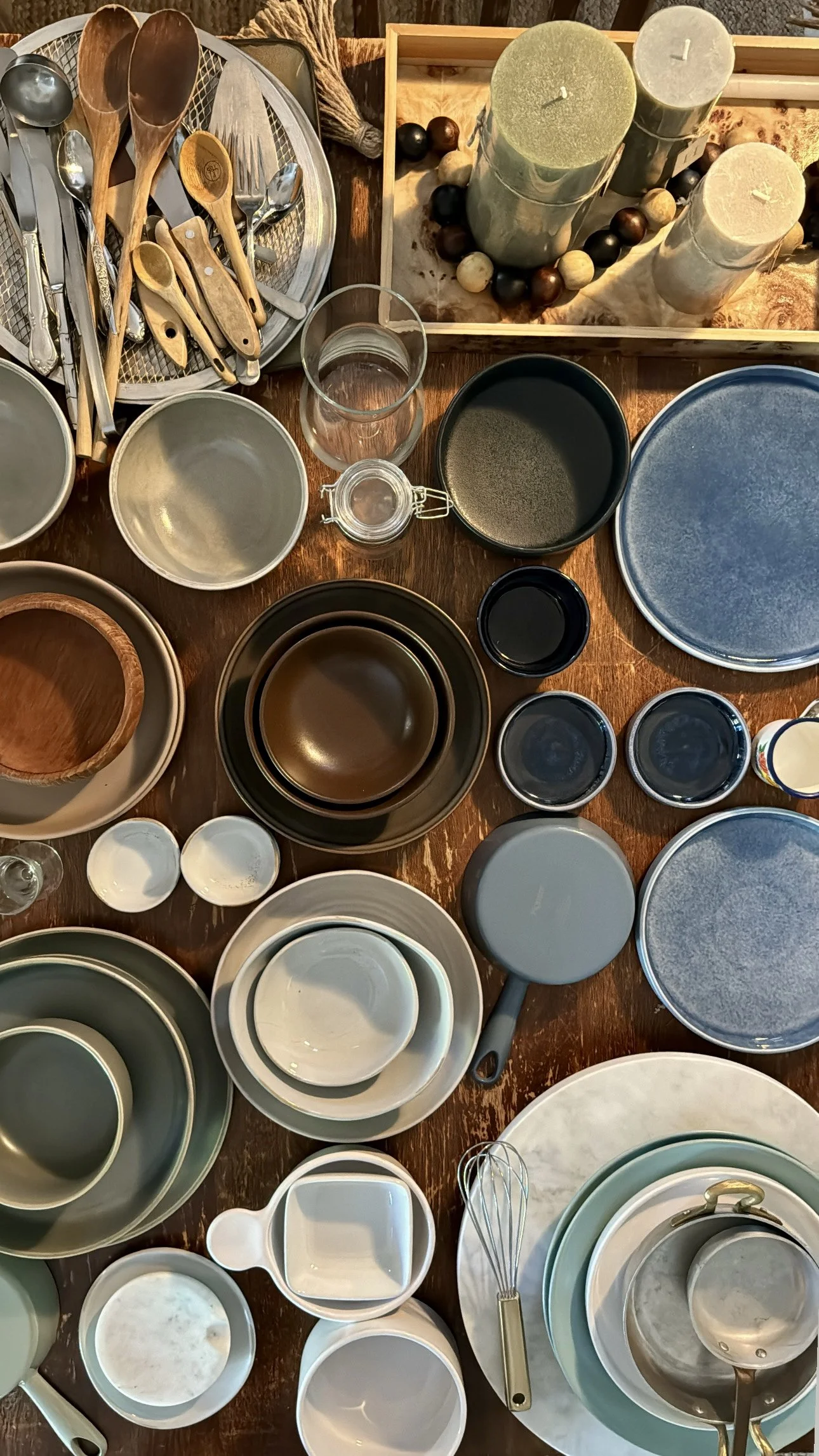 A collection of various kitchenware items including bowls, plates, cups, jars, candles, and utensils on a wooden table.