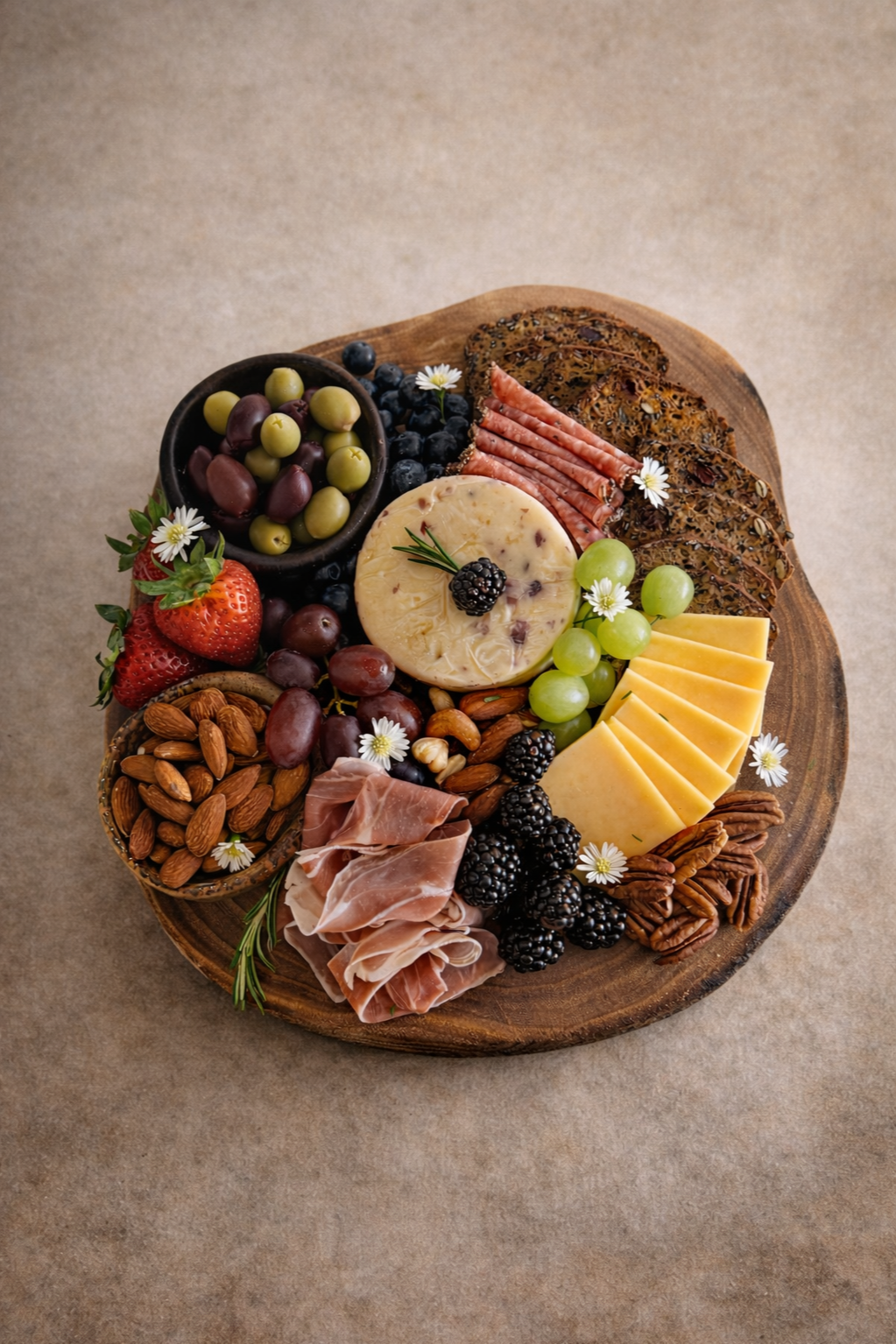 A round wooden cheese and charcuterie board with various cheeses, grapes, berries, nuts, cured meats, and crackers, decorated with small white flowers.