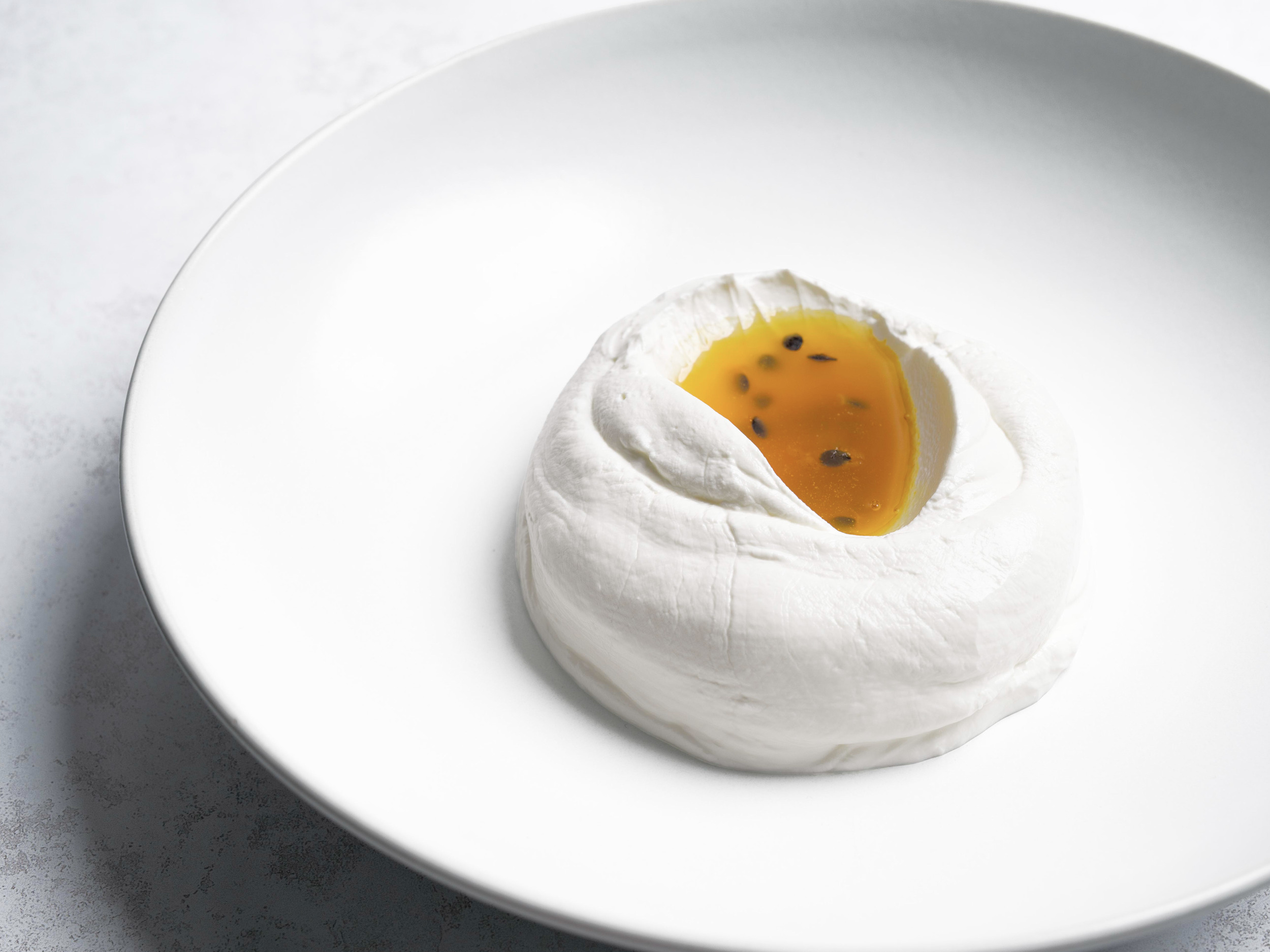A soft cheese with a cracked open top revealing a golden passion fruit filling with black seeds inside, served on a white plate.