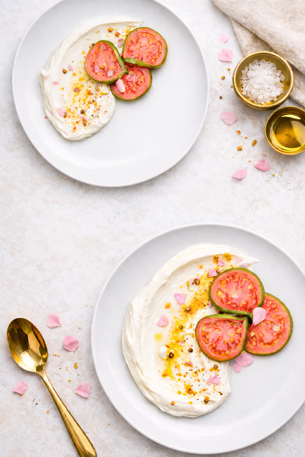 Two plates of burrata cheese topped with sliced tomatoes, pink flower petals, and drizzled with olive oil, served on a white surface with pink flower petals and small bowls of salt and oil nearby.