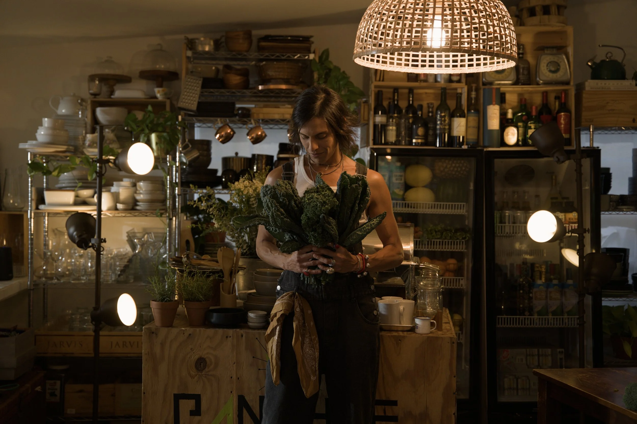 A woman in a sleeveless top holding a bunch of kale in a cozy kitchen with warm lighting, shelves stocked with bottles, cookware, and plants.
