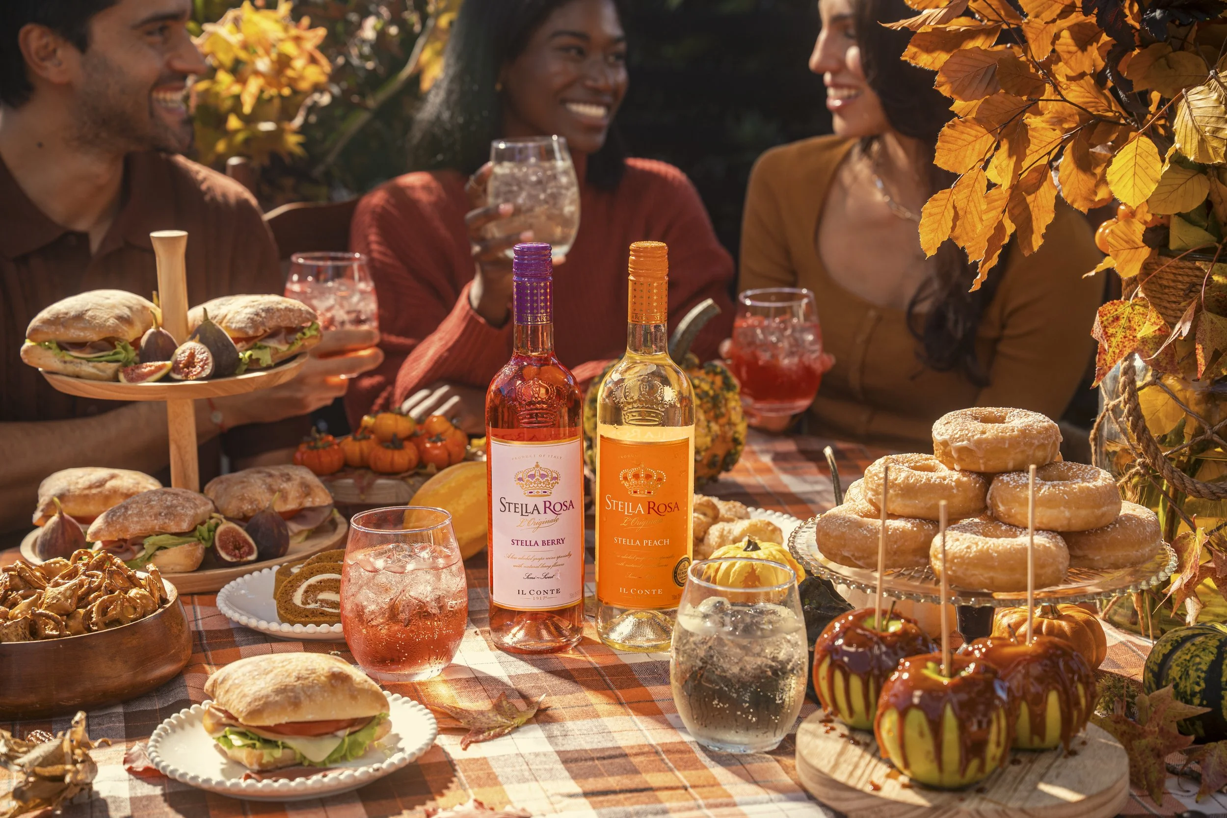 People enjoying an outdoor autumn picnic with fall foliage, wine bottles, and a variety of desserts and sandwiches on a checkered tablecloth.