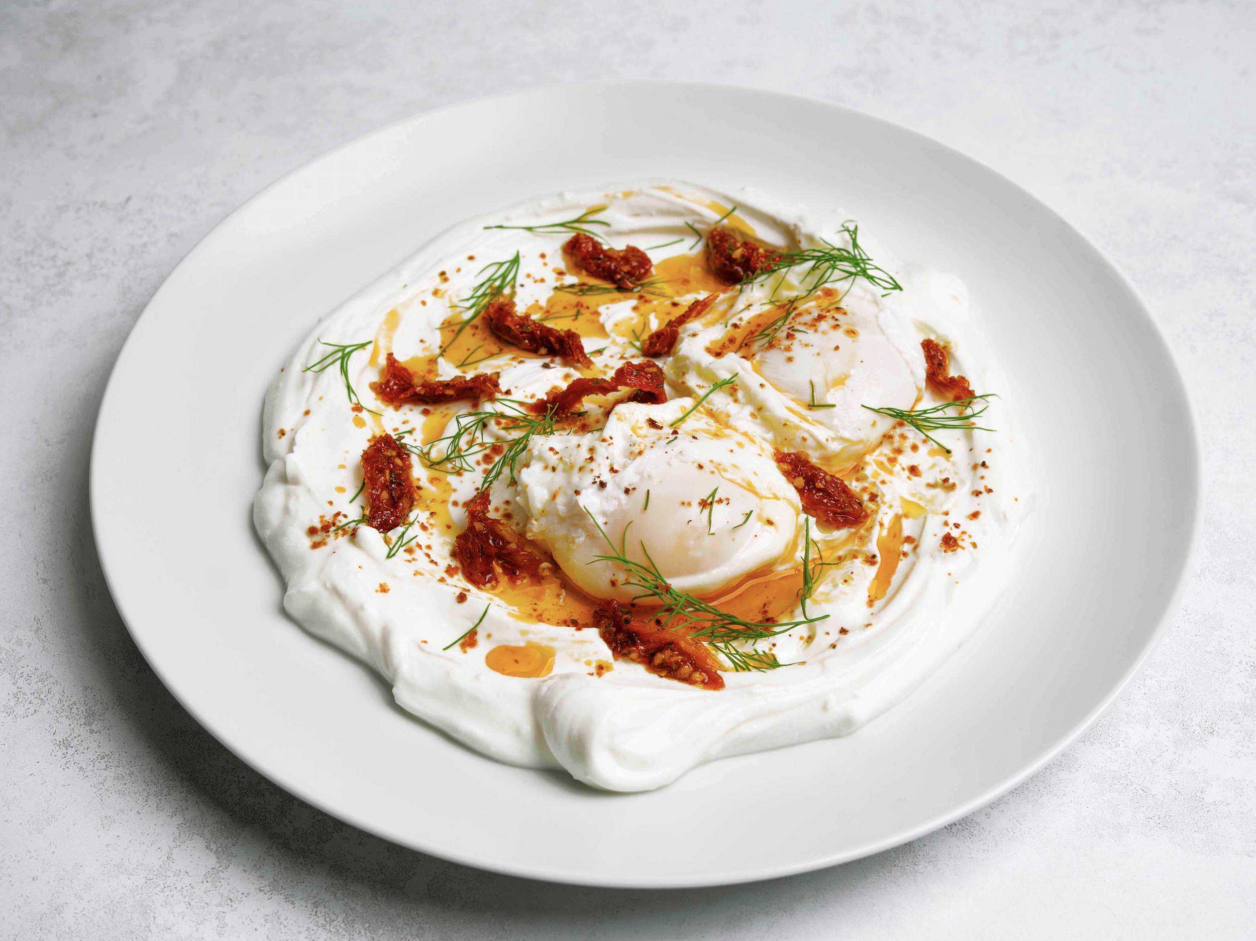 Poached eggs topped with sun-dried tomatoes, fresh dill, and drizzled with hot sauce on a white plate.