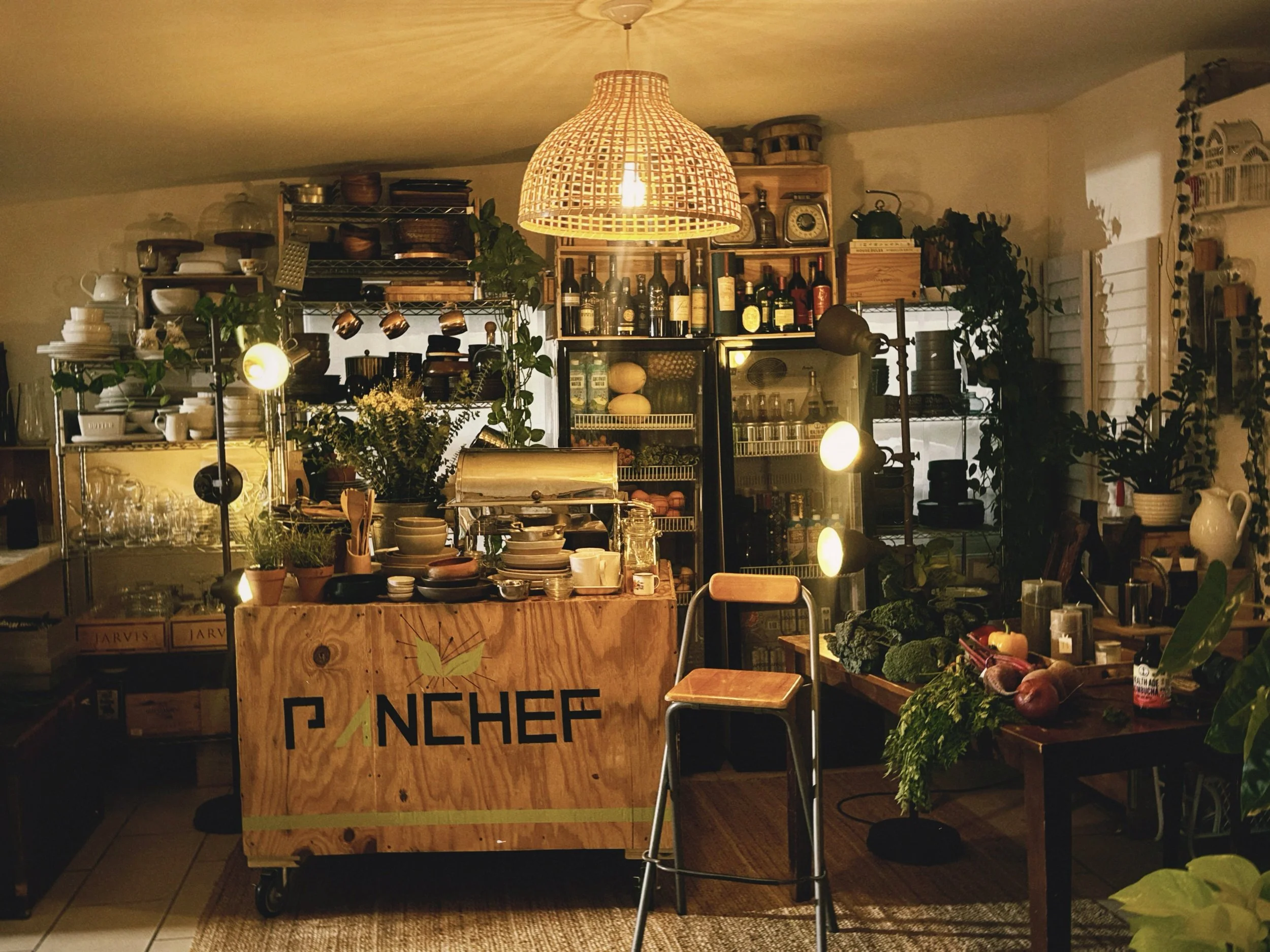 A cozy kitchen or cafe setting illuminated by a woven rattan pendant light. There are shelves filled with bottles, jars, and kitchenware in the background. The counter displays various bowls, plants, and fresh vegetables. A wooden sign with the words 'FRENCH' is visible in the foreground.