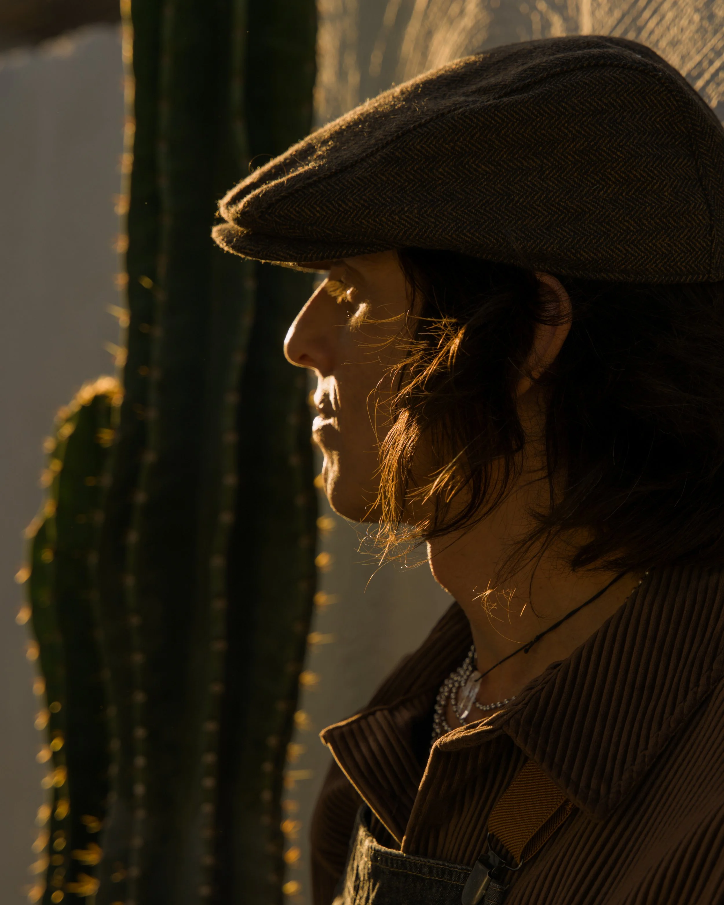 Side profile of a woman wearing a brown herringbone-pattern hat, a brown corduroy jacket, layered necklaces, and earrings, with sunlight highlighting her face and hair. There are tall cacti in the background.